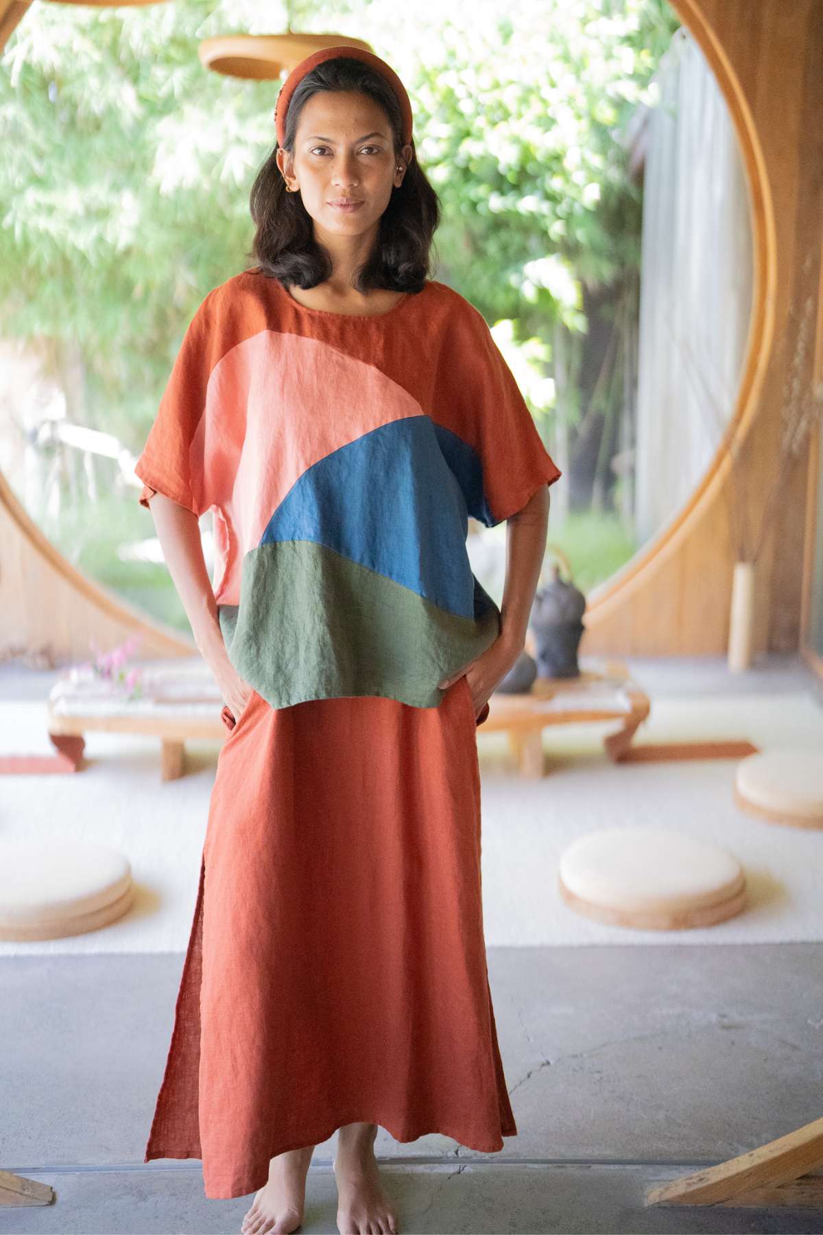 In a tranquil room featuring a circular window that offers a view of the garden, someone stands wearing the Myrah Penaloza Uluwatu Top, an orange garment with a vibrant pattern that exudes relaxation and poise. The light illuminates the minimalist decor, adorned with floor cushions and emphasizing sustainability through its natural elements.