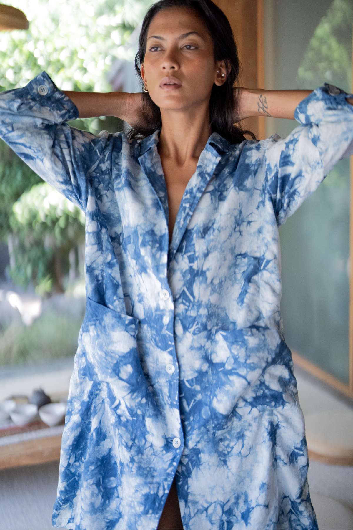 A person stands with their arms behind their head, wearing the Rubi Linen Jacket Indigo by Myrah Penaloza. The background features blurred greenery and a softly lit interior space, perfectly complementing the relaxed mood.