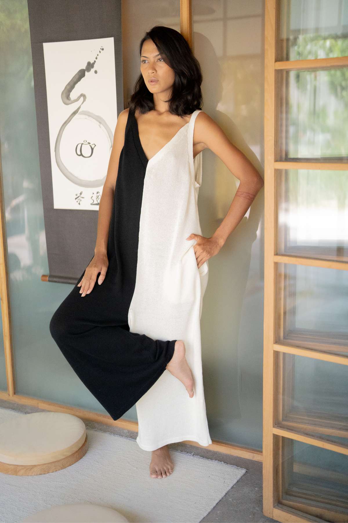 In a minimalist room adorned with wooden sliding doors, a person stands wearing the Yin Yang Kundalini Knitted Playsuit from Myrah Penaloza. They casually lean against the wall next to an abstract art scroll, bathed in soft lighting that complements the tranquil ambiance reminiscent of cotton knit wide-leg bottoms.