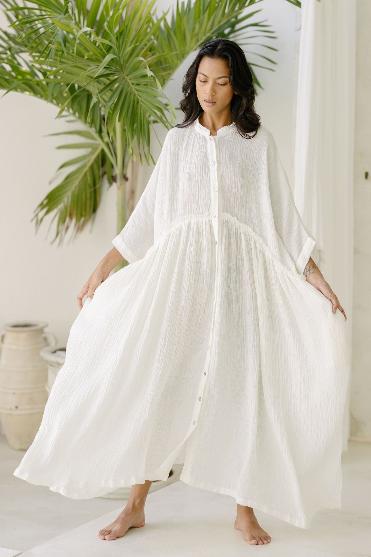 A woman in a white, flowing Myrah Penaloza Bona Dress (Cotton/Linen Crinkle) stands barefoot in a minimalist, airy setting with light-colored walls and a potted palm plant in the background. The dress features long sleeves, buttons down the front, and a loose, billowing silhouette—perfect for holiday season events.