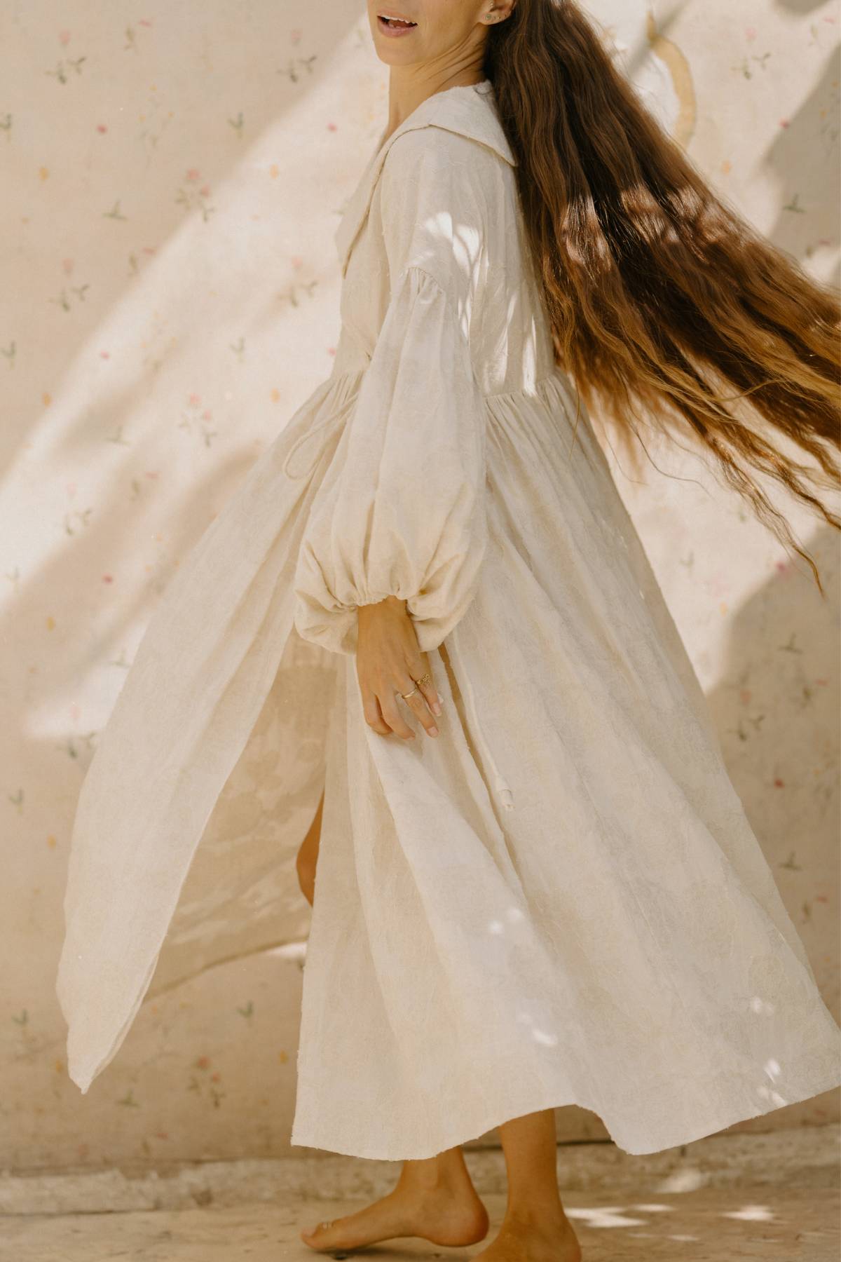 A person with long brown hair is standing barefoot on a textured surface, wearing the Blooming Magdalena Dress by Myrah Penaloza. The dress boasts billowy sleeves and has a slight opening, while the background showcases a soft floral pattern.