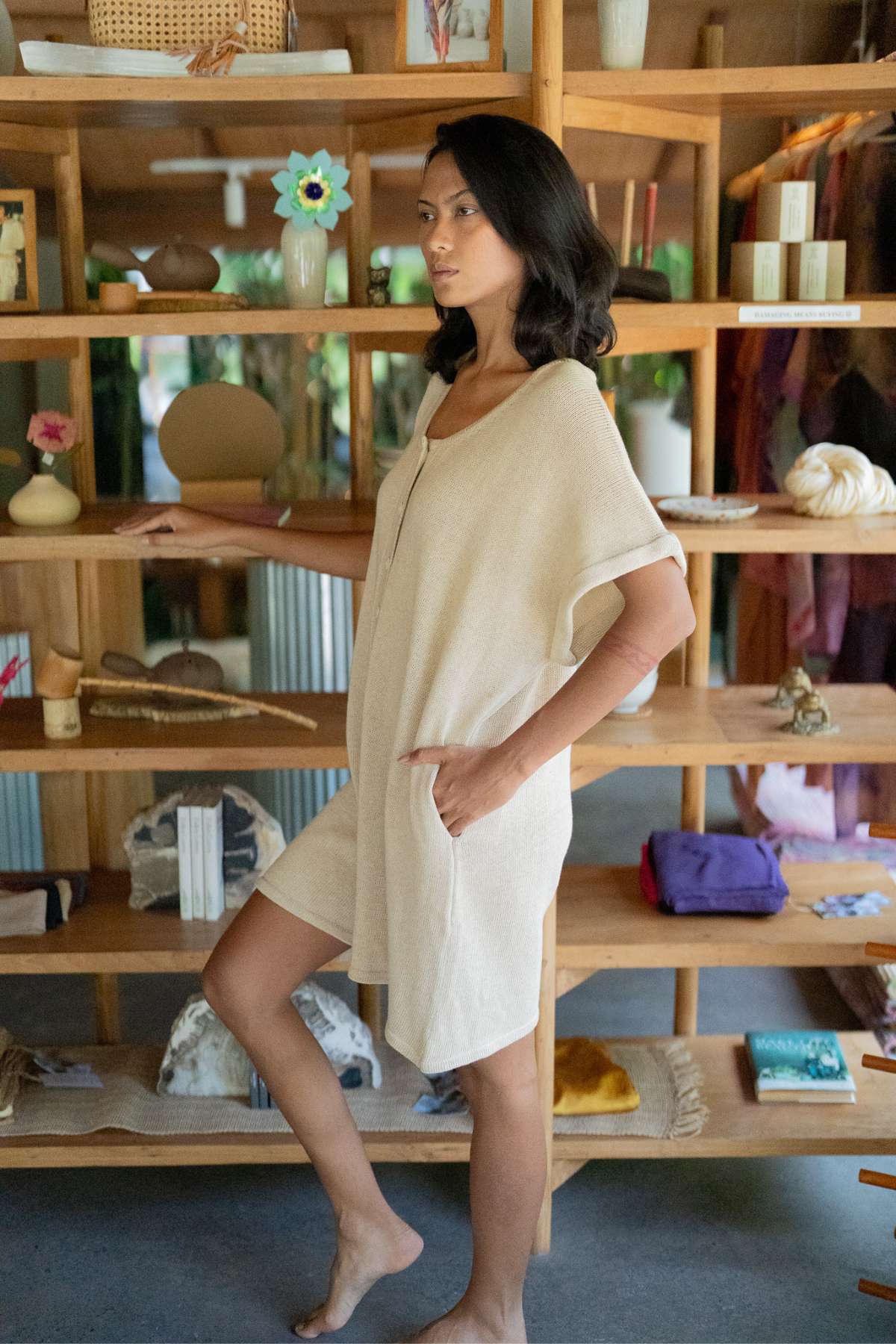 A woman in a beige Henley Knitted Playsuit from Myrah Penaloza stands barefoot beside a wooden shelf filled with books, decor, and fabric. The room exudes a natural, serene atmosphere with an open window in the background.