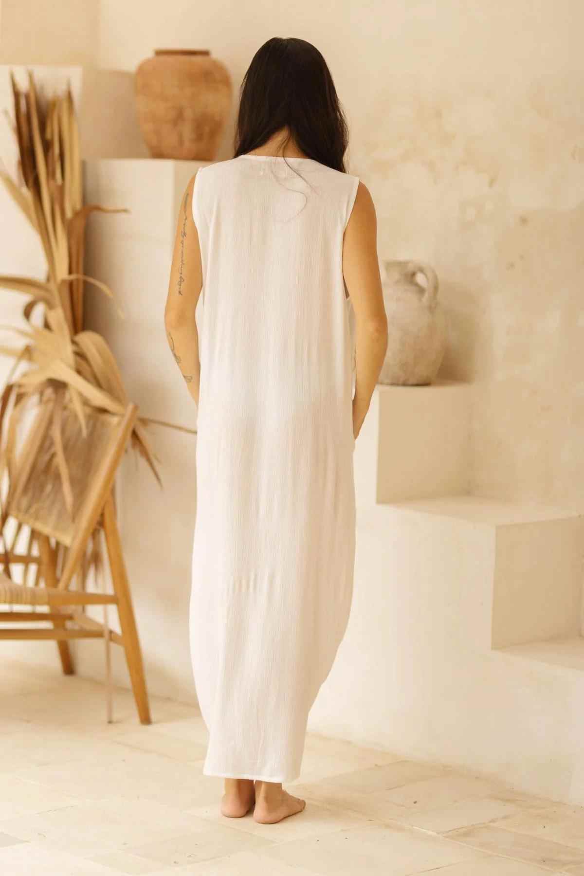 A person with long dark hair is seen from the back wearing the Virgo Luna Dress by Myrah Penaloza. The sleeveless, high neckline floor-length white dress drapes elegantly as they stand barefoot on a light-colored surface, surrounded by neutral-toned decorative elements and dried plants in the background.