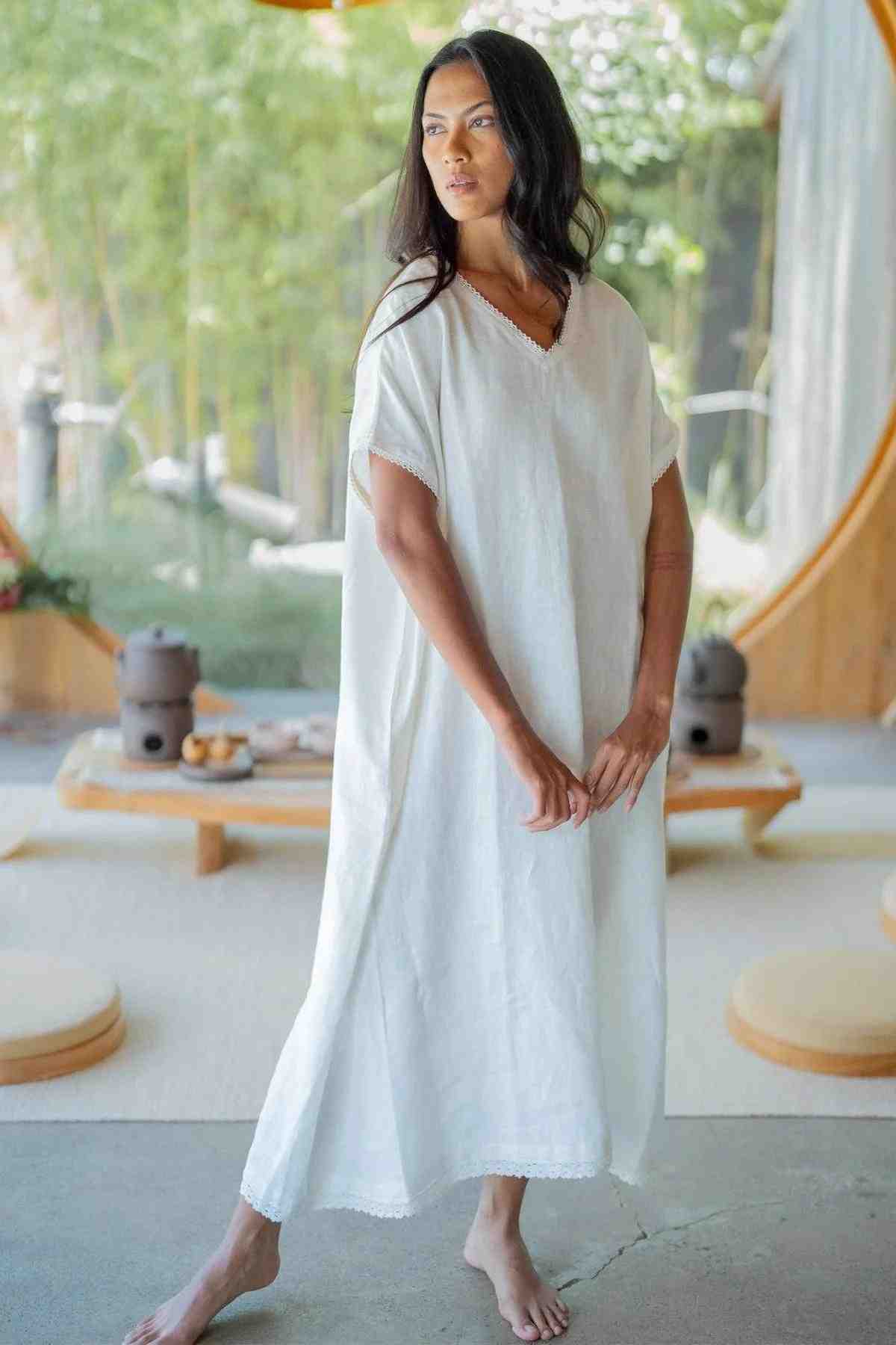 A barefoot person with long dark hair and a thoughtful expression stands indoors, wearing the Ruby Lace Tunic by Myrah Penaloza— a loose, white calf-length dress with a V neckline. The background features a cozy, modern room with wooden accents, low seating, and greenery visible through large windows.