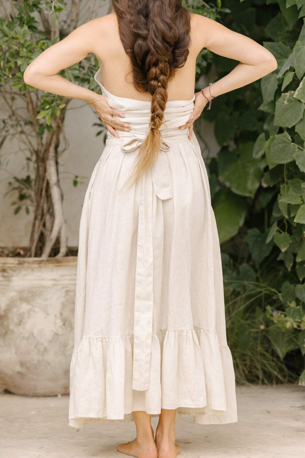 A woman with long braided hair stands barefoot outdoors, facing away. She wears a flowing, strapless light flax dress with a tied waist that echoes the elegant grace of Myrah Penaloza's Viva Skirt. Green foliage and a large potted plant enhance the serene setting.