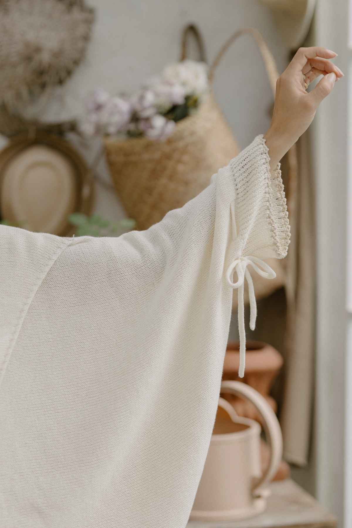 A close-up of an arm in the Honey Knit Sweater by Myrah Penaloza, featuring a loose-fitting, cream-colored sleeve adorned with a tied bow at the wrist. The background showcases a cozy, rustic scene with blurred objects such as a woven basket, dinner plates, and a watering can.