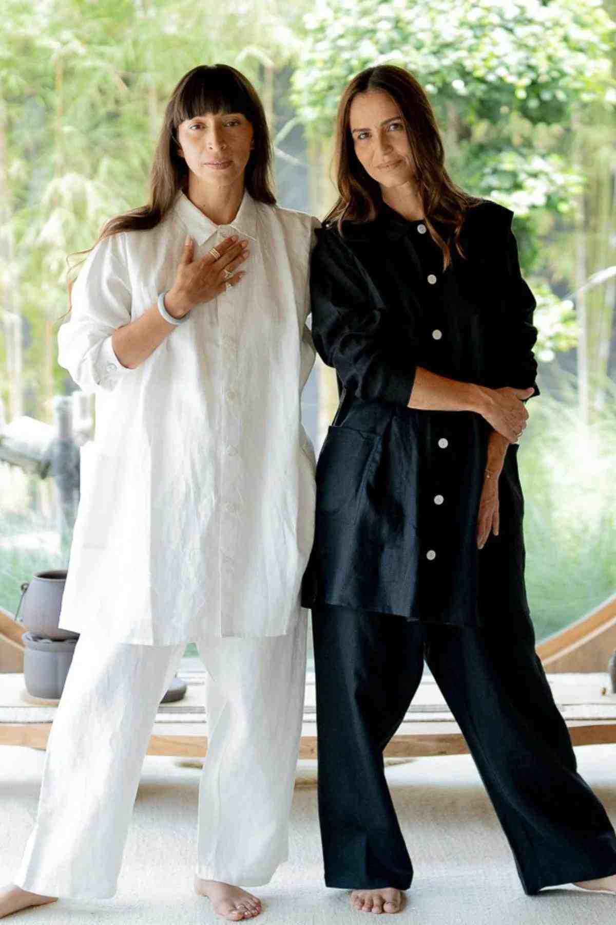 Two women stand side by side indoors, both with long brown hair and barefoot. The woman on the left wears an eclectic fashion ensemble featuring a loose white outfit, while the woman on the right dons a deep royal blue Tender Jacket (100% Linen) from Myrah Penaloza. Each has one hand gently resting on their chest, with greenery visible in the background.