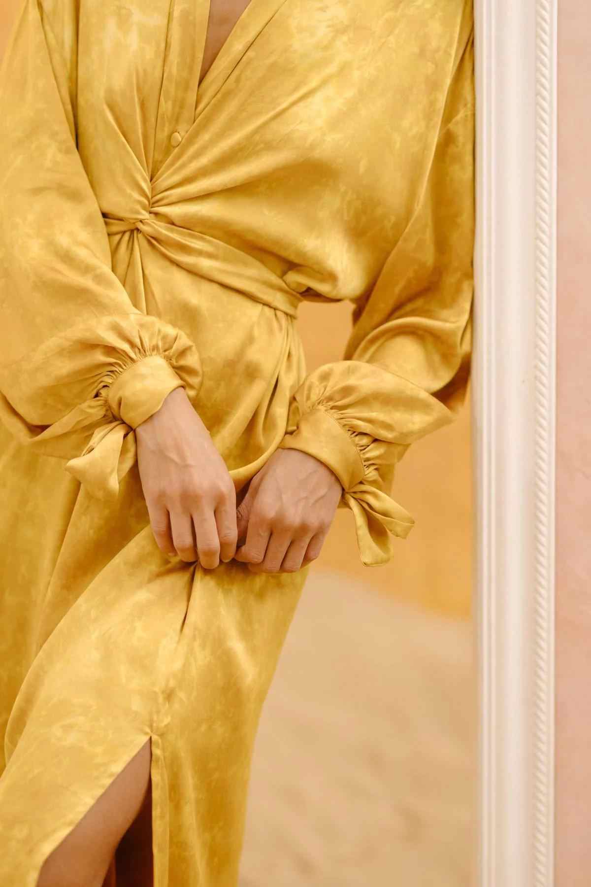 A person wearing a long-sleeved, mustard yellow Silk Inanna Wrap Gown by Myrah Penaloza, featuring a deep V-neck and gathered cuffs, stands near a white door frame. This exquisite dress is crafted by artisan tailors from botanically hand-dyed silk and includes a waist tie and a side slit that reveals part of the leg. The background is filled with soft, warm tones.