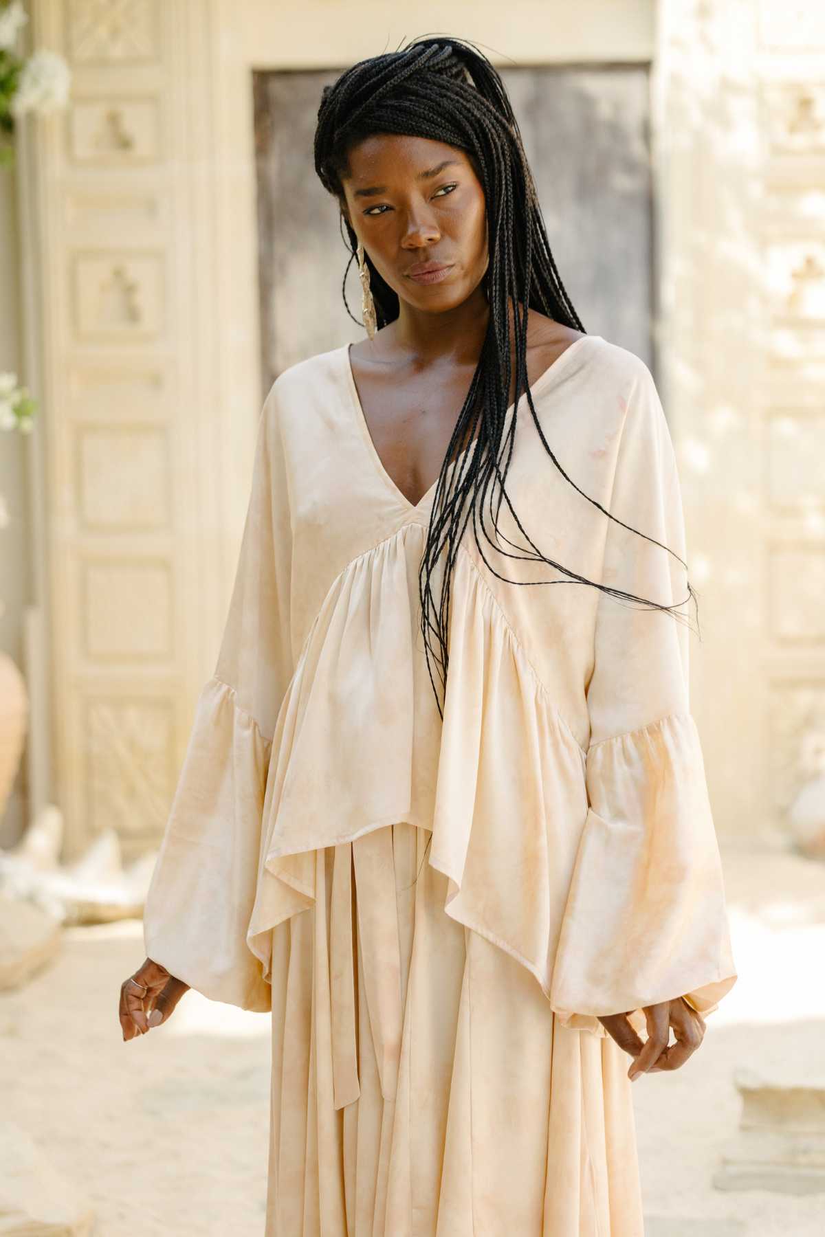 A woman stands outdoors wearing the elegant Eva Top & Orisa Dance Skirt Set, crafted from botanically hand-dyed silk by Myrah Penaloza. Her long braided hair complements the light-colored Mediterranean architectural setting, adding elegance and charm to the serene scene.