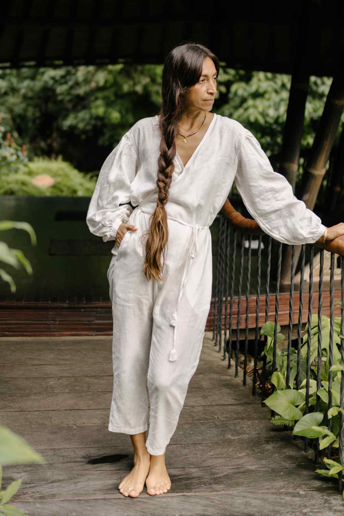 A woman with long, braided hair stands barefoot on a wooden deck amidst lush greenery. She is dressed in Myrah Penaloza's Linen Long Sleeve Dora Playsuit, showcasing its unique detailing and adjustable waistline for an elegant simplicity. She leans slightly against the railing, gazing contemplatively into the distance.