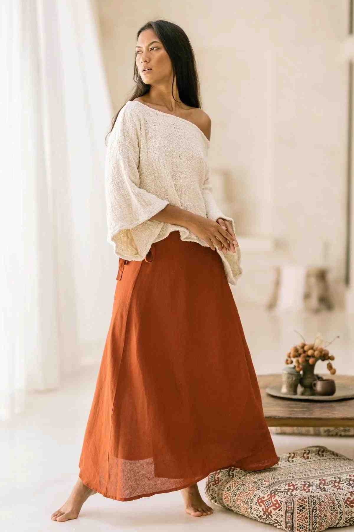 A woman stands barefoot in a minimalistic room with white walls and soft lighting, dressed in the Spirit Top & Shanti Wrap Skirt Set from Myrah Penaloza. The loose, off-the-shoulder white top pairs elegantly with the long, adjustable burnt orange skirt made of 100% linen. She gazes serenely to the side, surrounded by woven baskets and a small table.