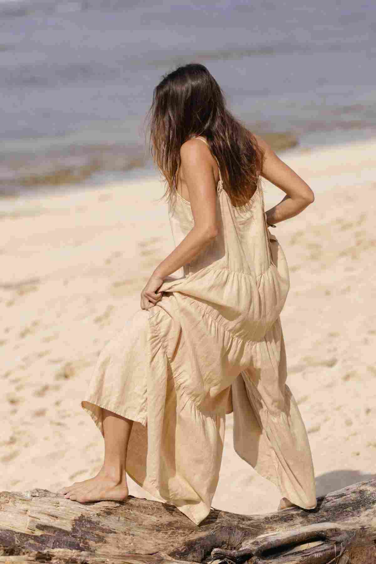 A woman wearing the Myrah Penaloza La Mexicana Playsuit, which is made to order from 100% linen, sits on a driftwood log at a sandy beach with adjustable straps flowing elegantly. Facing away from the camera, she gazes at the ocean under a clear sky, creating an idyllic scene of effortless resort wear elegance.