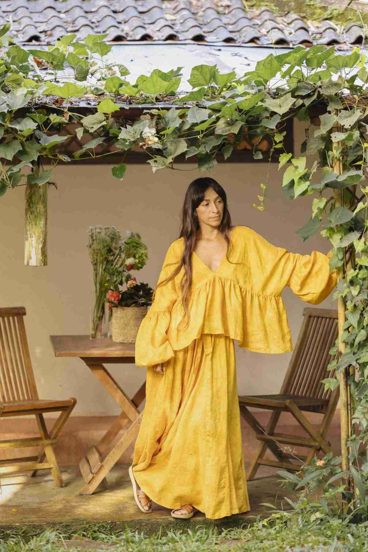 A woman wearing a flowing, bright yellow Eva Linen Top with flowering sleeves by Myrah Penaloza stands near the doorway of a wooden patio covered with green foliage. Behind her is a small table with two wooden chairs and a vase of flowers. The atmosphere is serene and garden-like.