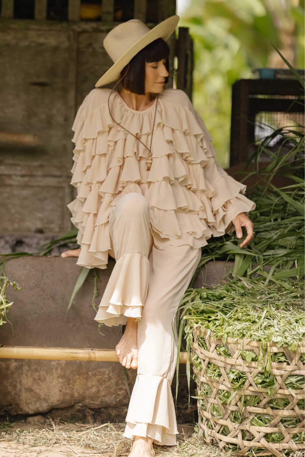 A woman with short hair sits in an outdoor setting, adorned in nature-inspired couture—a beige, ruffled outfit from Myrah Penaloza's Anusara Bloom - Made To Order collection, paired with a wide-brimmed hat. Barefoot and relaxed with one leg propped on a ledge, she is beside a basket filled with green foliage. The rustic background complements the limited edition look.