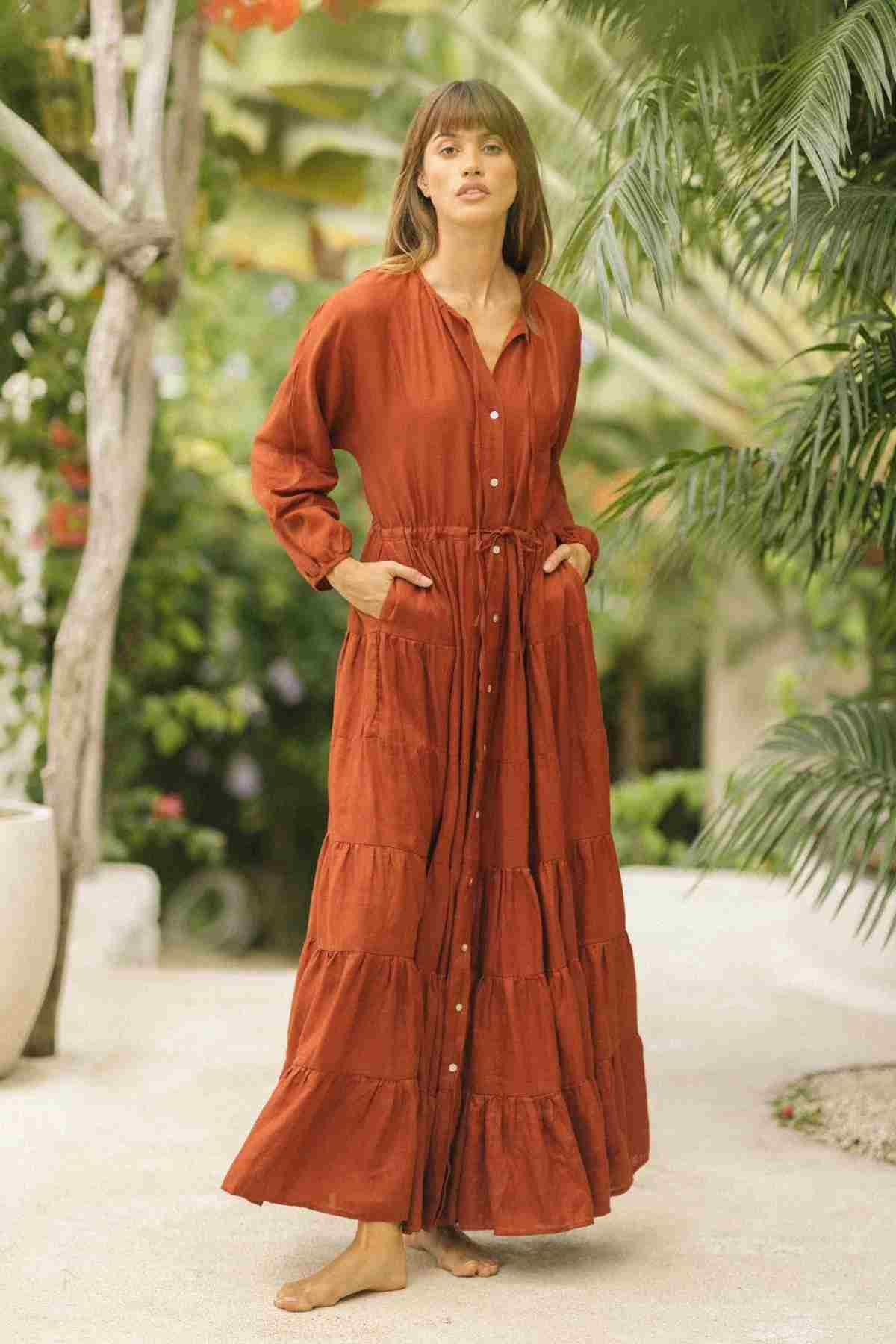 Dressed in a Myrah Penaloza La Mexicana Gown, the woman moves gracefully along a pathway enveloped by verdant foliage. The gown's rust hue complements the sunny, tropical setting as she looks ahead, her hands casually tucked into its adjustable waist pockets.