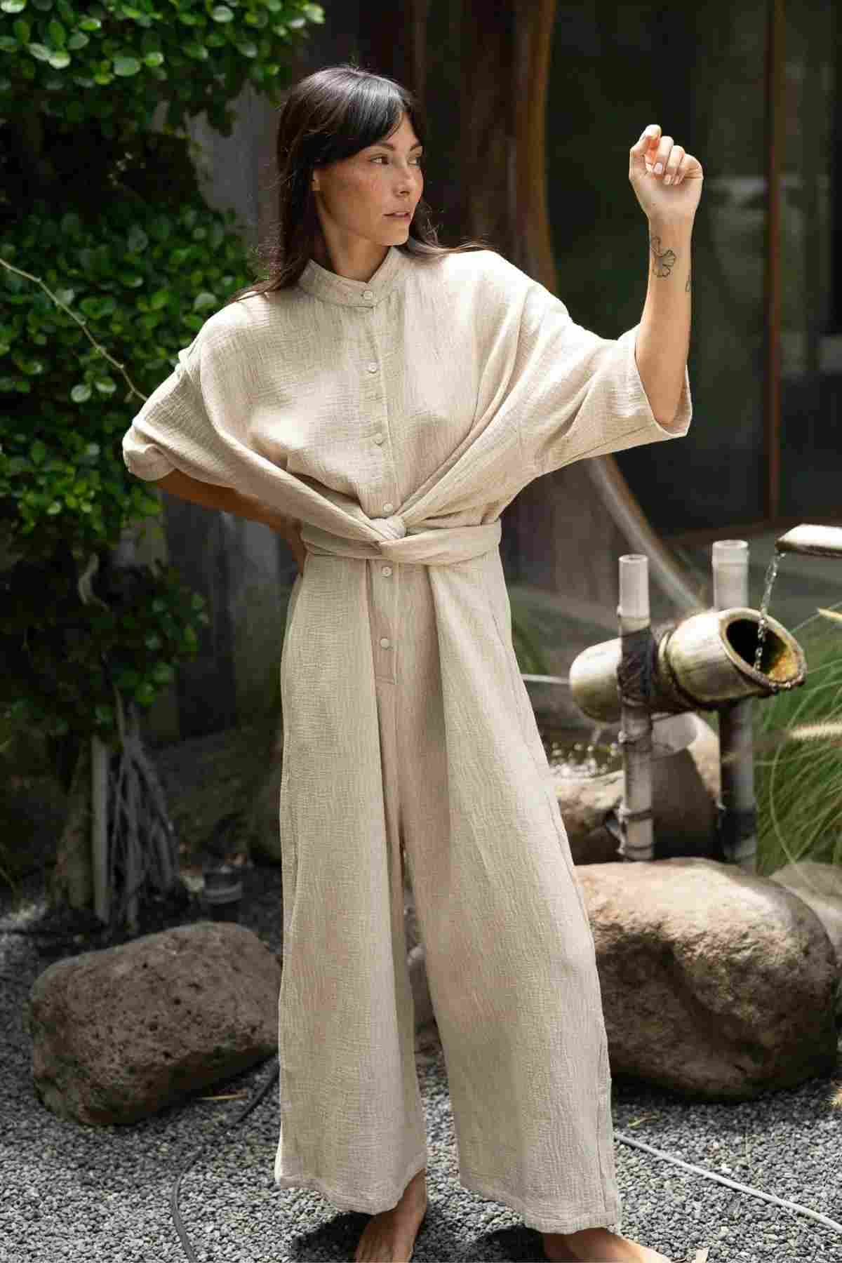 A woman with long, dark hair stands outdoors on a gravel surface, flaunting the Pine Playsuit by Myrah Penaloza. Made from a light beige cotton linen blend and featuring long sleeves, this versatile jumpsuit is showcased as she strikes a pose with her left hand on her hip and her right arm raised, revealing a small tattoo on her wrist. Lush greenery and rocks provide the perfect backdrop for this stylish wardrobe addition.