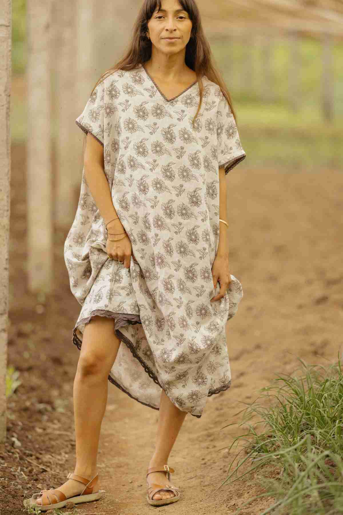 A woman stands in a natural outdoor setting, wearing the Ruby Lace Tunic by Myrah Penaloza. This loose, floral-patterned tunic features a V neckline and complements her long dark hair as she stands barefoot on the dirt path. The background is blurred, highlighting trees and greenery, creating a serene and earthy ambiance.