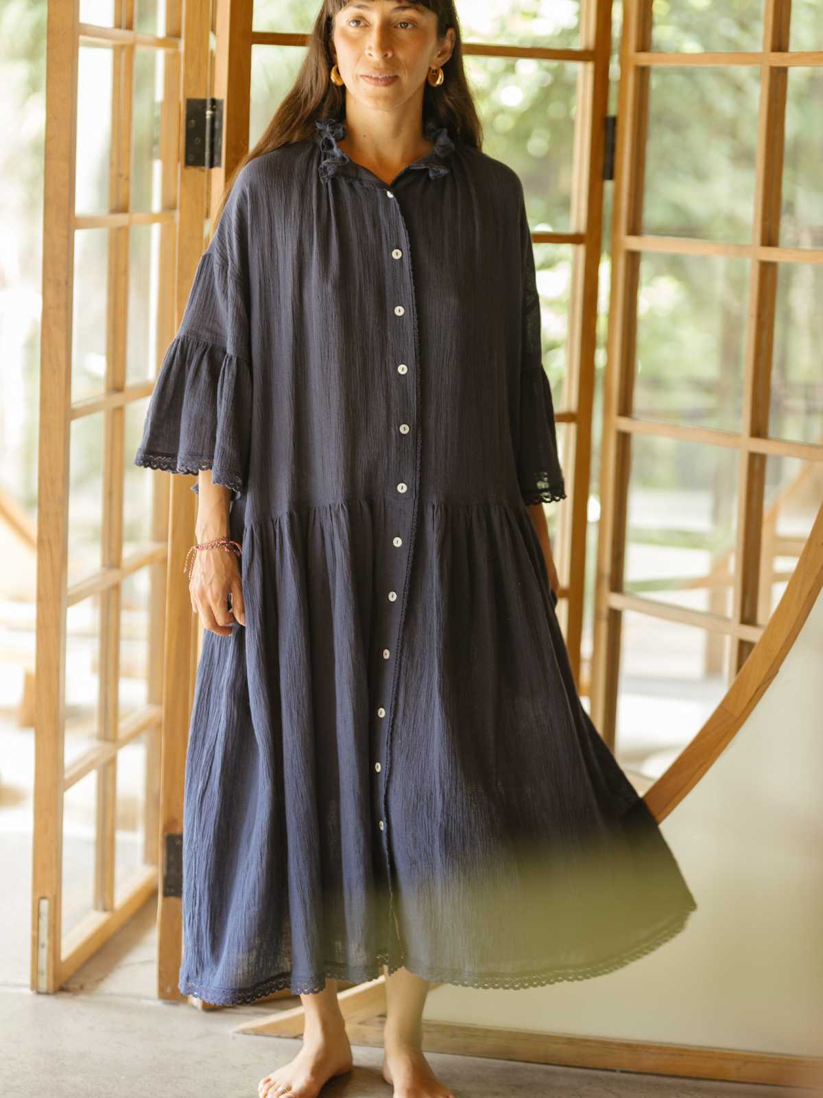 A woman stands barefoot in a doorway, adorned in a flowy, dark blue Juliette Linen Gown with ruffled sleeves and collar. The serene atmosphere is enhanced by the backdrop of wooden doors and greenery, all crafted by the brand Myrah Penaloza.