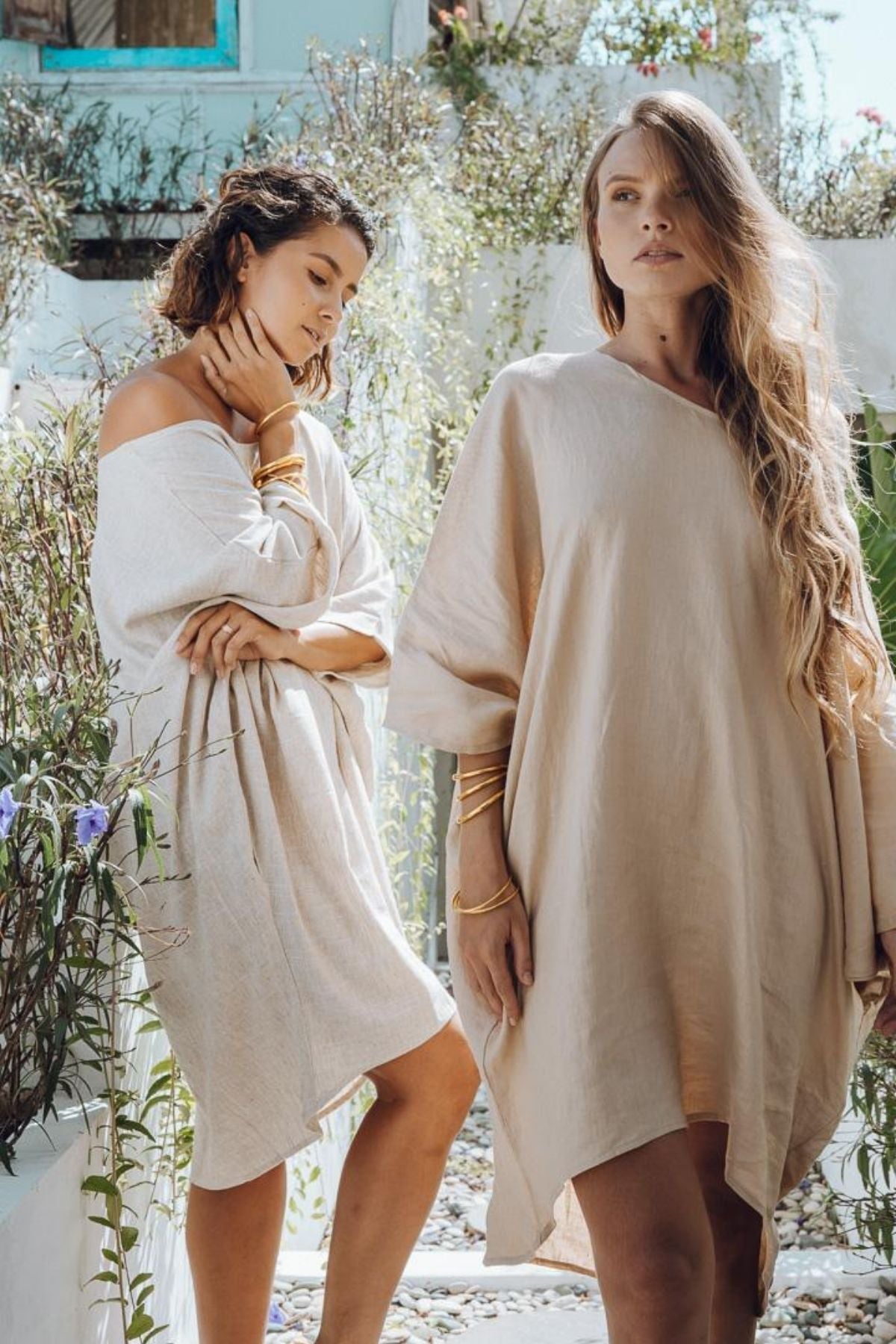 Two women dressed in loose-fitting Butterfly Tunics from Myrah Penaloza stand outdoors in a garden setting. One has her hand on her chin, while the other gazes forward, surrounded by lush greenery and flowers that create a serene atmosphere.