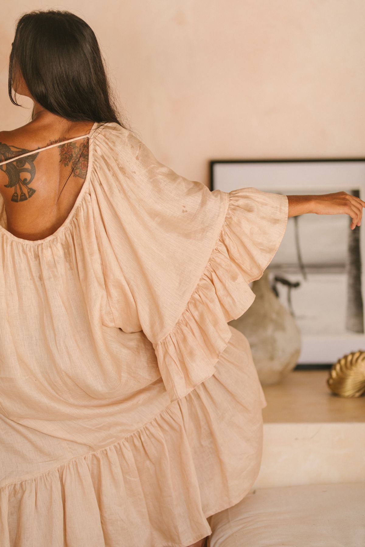 A woman with long dark hair and a shoulder tattoo wears a flowing Brida Linen Gown Long by Myrah Penaloza. She stands in a minimalist room, where a framed picture and decor rest on the shelf. Each element, including her dress, is chosen with care for elegance and carbon-neutral shipping.