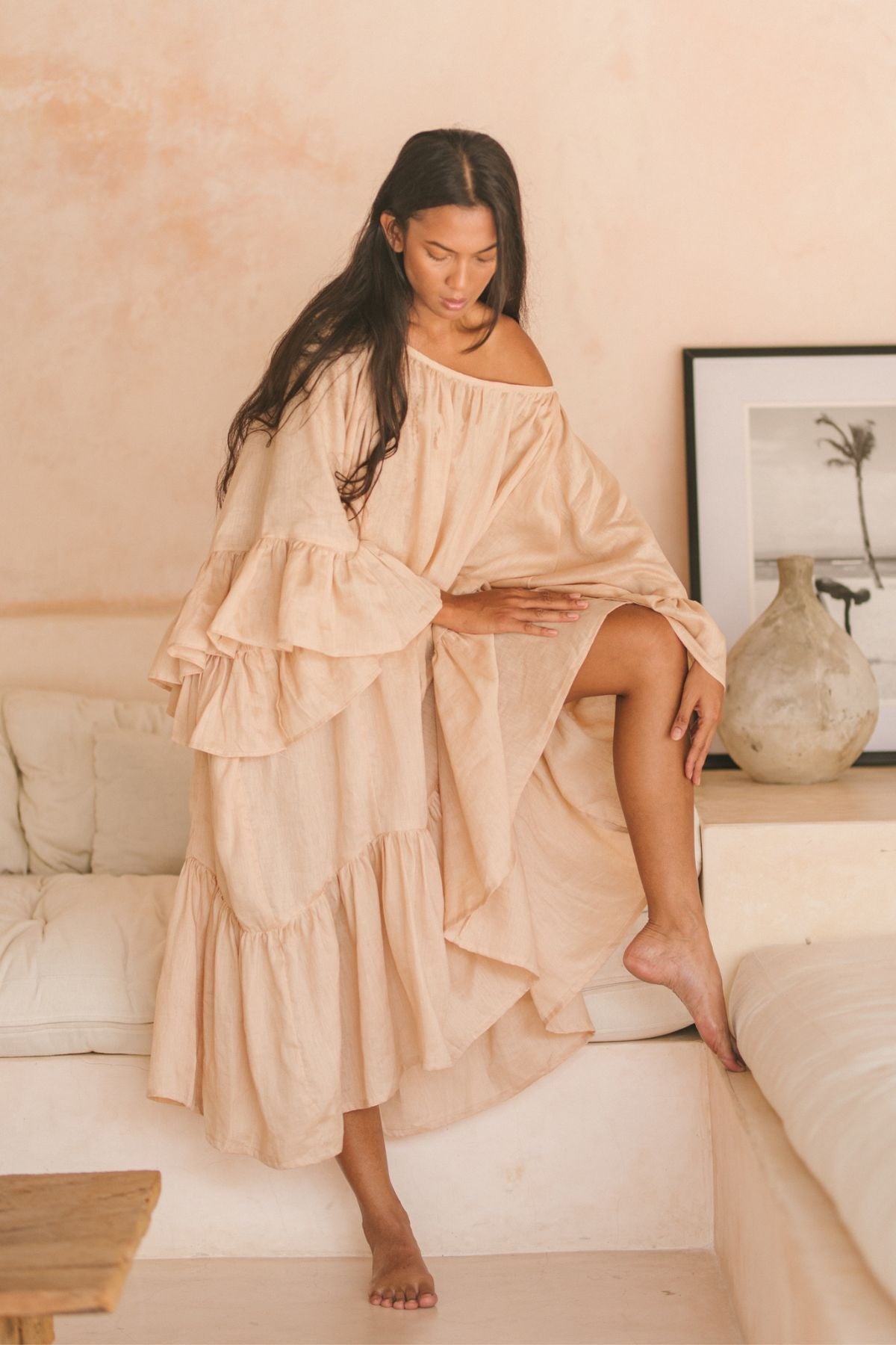 A woman in a flowing Brida Linen Gown Long by Myrah Penaloza stands barefoot on a step in a softly lit room, her long hair cascading over her shoulders. The background features a light-colored sofa, a vase, and a framed monochrome photograph.