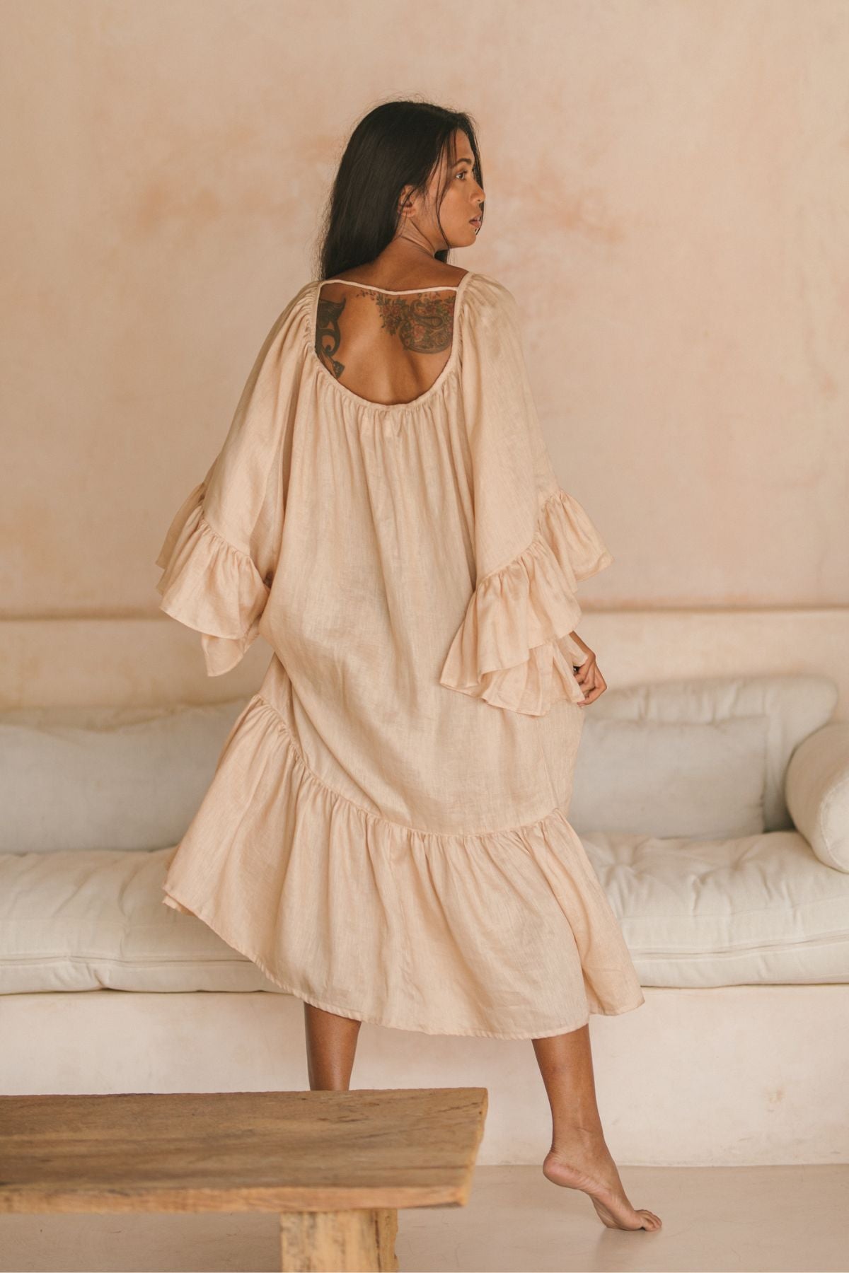 A woman with long hair is barefoot in a room with a light beige wall, wearing a Myrah Penaloza Brida Linen Gown Long. The light pink dress has ruffled sleeves and hem, showing her shoulder tattoo. A white sofa and wooden table complete the serene setting.