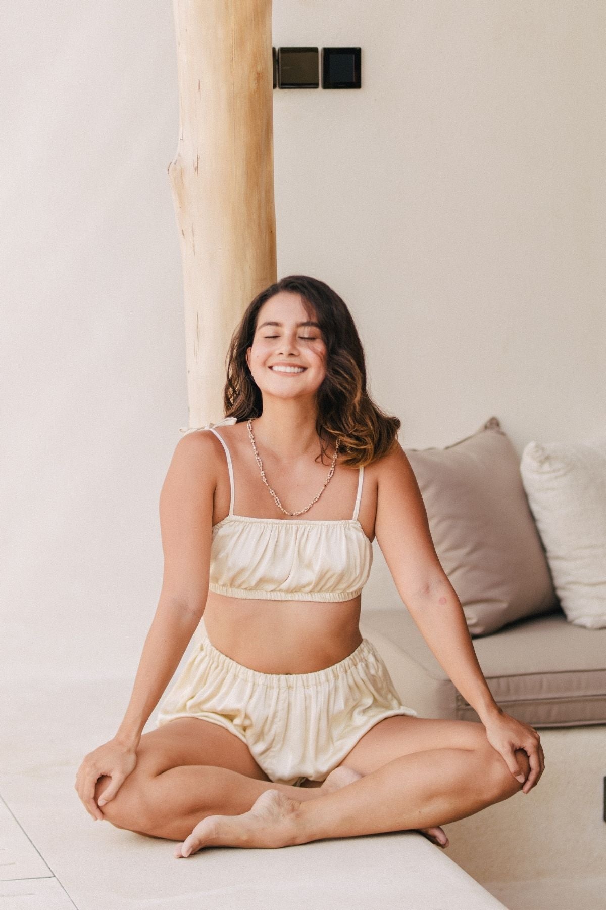 A woman with shoulder-length hair is sitting cross-legged on the floor, smiling with her eyes closed. She is wearing the Lucid Dreamer Underwear Set by Myrah Penaloza, a cream-colored satin two-piece outfit consisting of a crop top and shorts. Behind her, there is a beige cushion and a wooden post, softly illuminated by the moonlight.