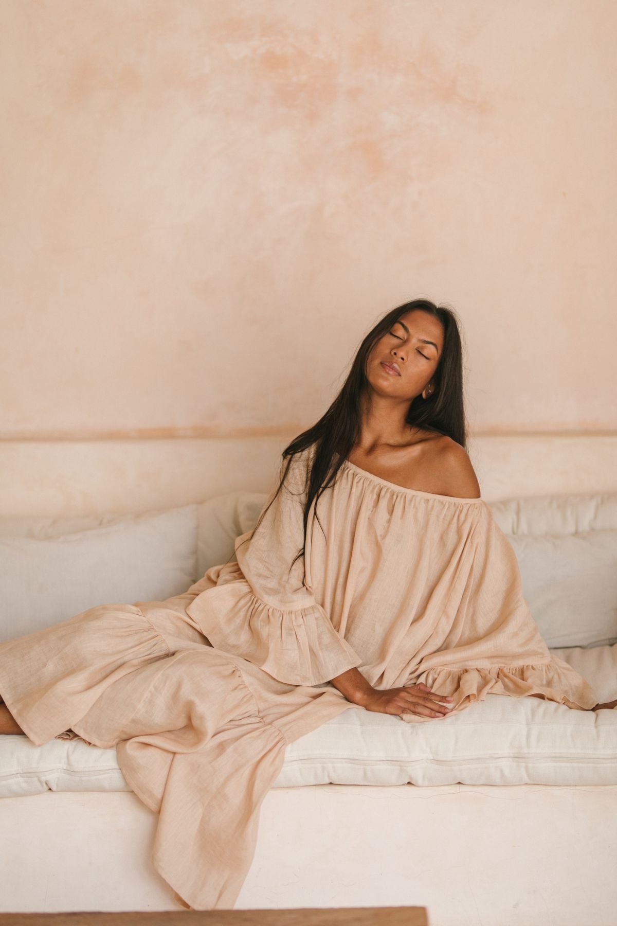 A woman in a flowing Brida Linen Gown Long by Myrah Penaloza sits on a white sofa against a peach-toned wall. Her long dark hair cascades over her shoulders, and with closed eyes, she appears serene. The room's soft, calming ambiance perfectly complements the gown's gentle elegance.