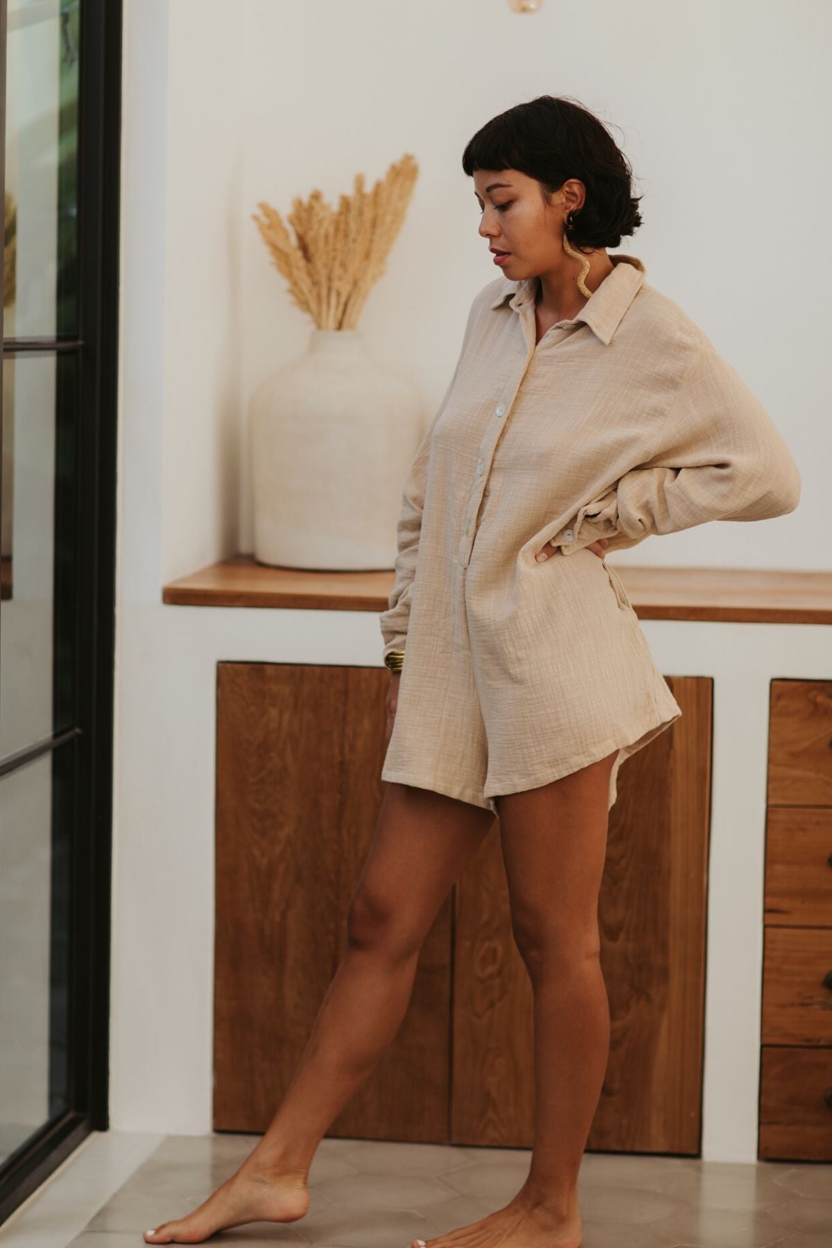 A barefoot woman with short dark hair stands indoors by a glass door, posing thoughtfully. She wears The Dreamer Playsuit in Spring Clay, crafted from 100% cotton and designed by Myrah Penaloza, and has one hand on her hip. A large vase with dried wheat is on the counter behind her.