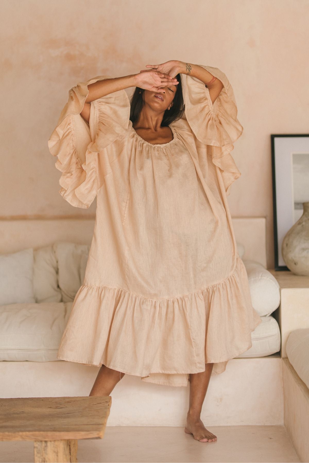 A person in a flowing Brida Linen Gown Long by Myrah Penaloza stands gracefully in a minimalist room with cream walls. One arm is raised above their head, while the other partially obscures their face. In the background, a couch and tasteful decor enhance the serene atmosphere.