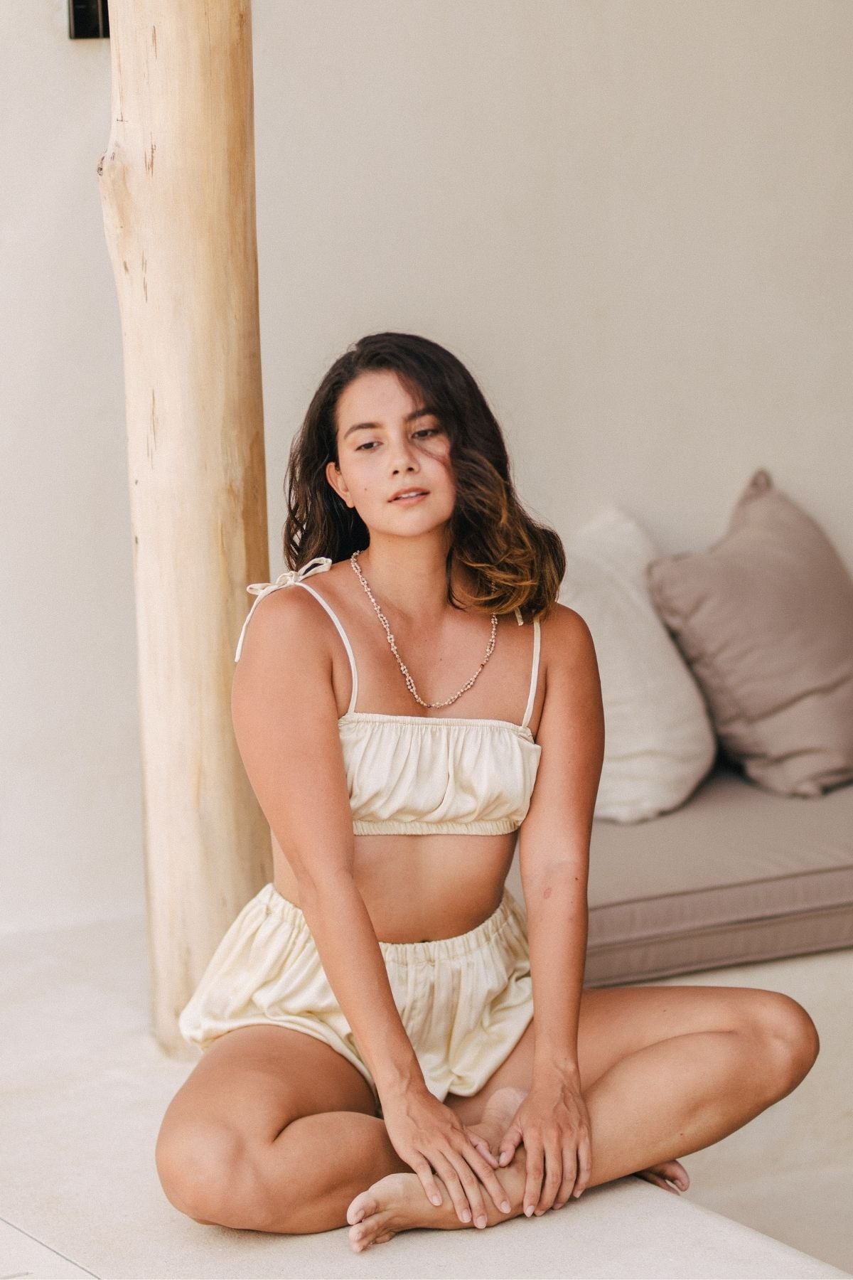 A woman with shoulder-length hair sits cross-legged on the floor, wearing the Lucid Dreamer Underwear Set (Silk/Satin) by Myrah Penaloza. This elegant beige two-piece outfit features a sleeveless top and matching shorts that resemble an upscale underwear set. Behind her, a wooden pillar and a sofa with beige cushions contribute to the relaxed and minimalistic setting.