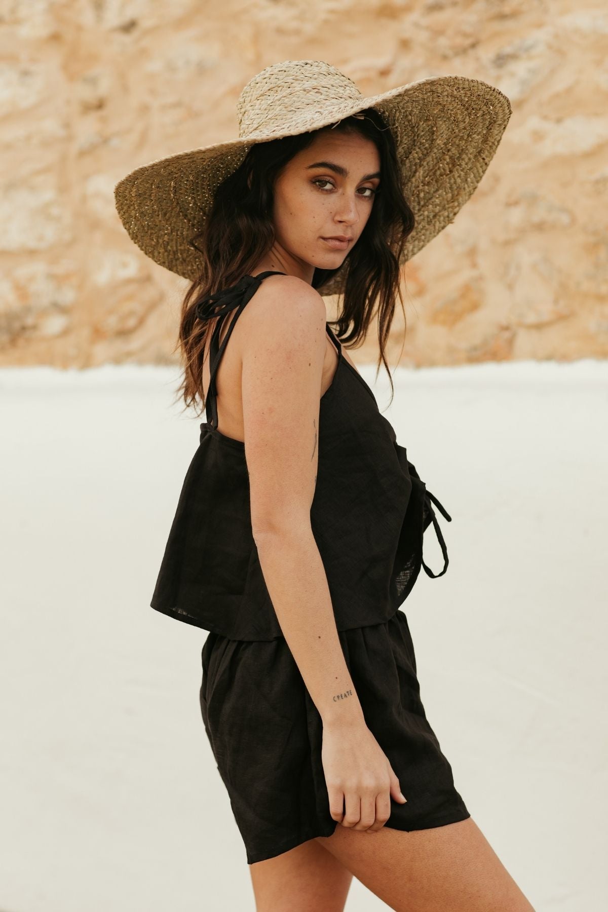 A woman with long dark hair is wearing a large straw hat and a black Nidra Sleeper Top by Myrah Penaloza, featuring adjustable straps. She stands against a stone wall backdrop, looking over her shoulder towards the camera.