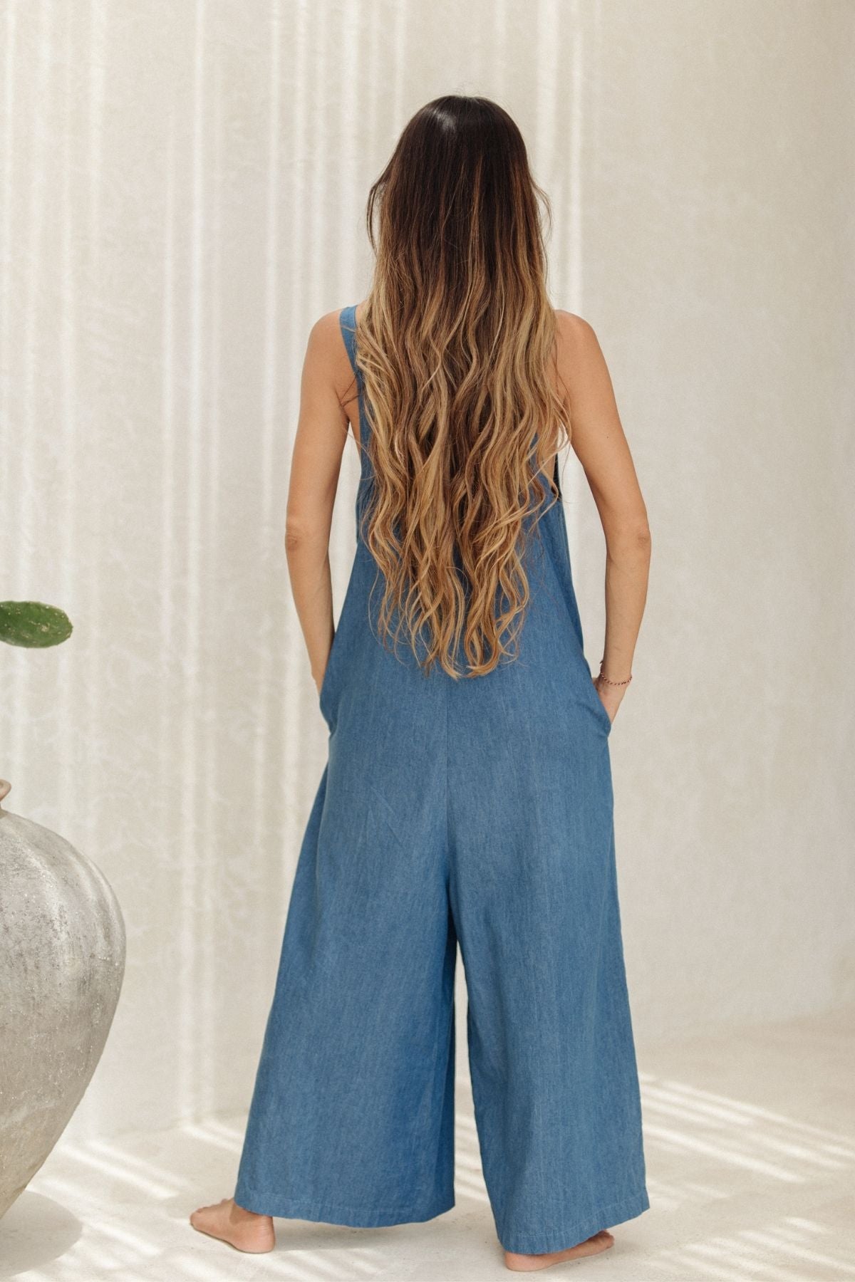 A person with long, wavy hair stands barefoot, facing away in a loose-fitting Denim Stonewashed Kundalini Playsuit by Myrah Penaloza. They are in a minimalistic, light-filled room with a large pot and subtly textured walls, evoking the serene ambiance of a Kundalini Yoga class.