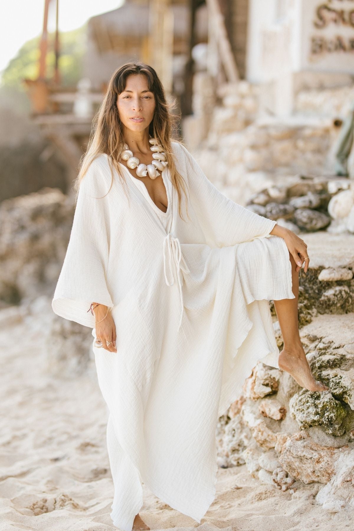 Wearing the Myrah Penaloza River Kaftan, made to order from 100% organic hemp and cotton, a person stands barefoot on a sandy beach, adorned with a large shell necklace. They lean against a rocky structure, with the ocean and greenery painting the serene backdrop.