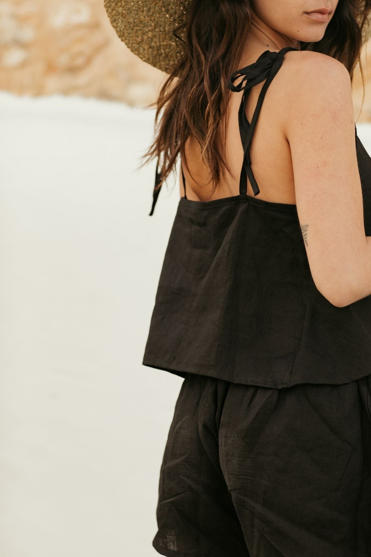 A woman wearing a straw hat and a black Nidra Sleeper Top by Myrah Penaloza stands outdoors. The background features a pale, sunlit landscape. Her hair falls over one shoulder, capturing the tranquil essence of Nidra yoga in this relaxed, summery scene.