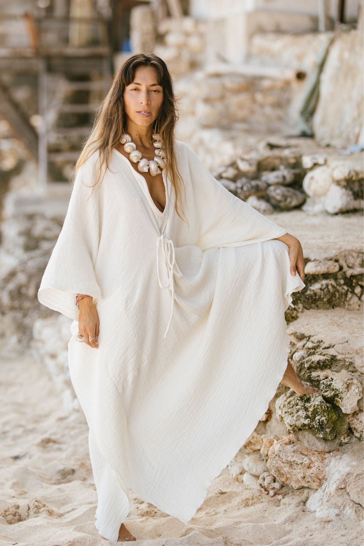 A woman stands on a sandy beach wearing the River Kaftan 100% Organic Hemp/Cotton by Myrah Penaloza, complemented with a chunky white necklace. She rests one foot on a rock step and gazes directly at the camera. The blurred stone structures in the background contribute to her earthy, carbon-neutral ensemble.