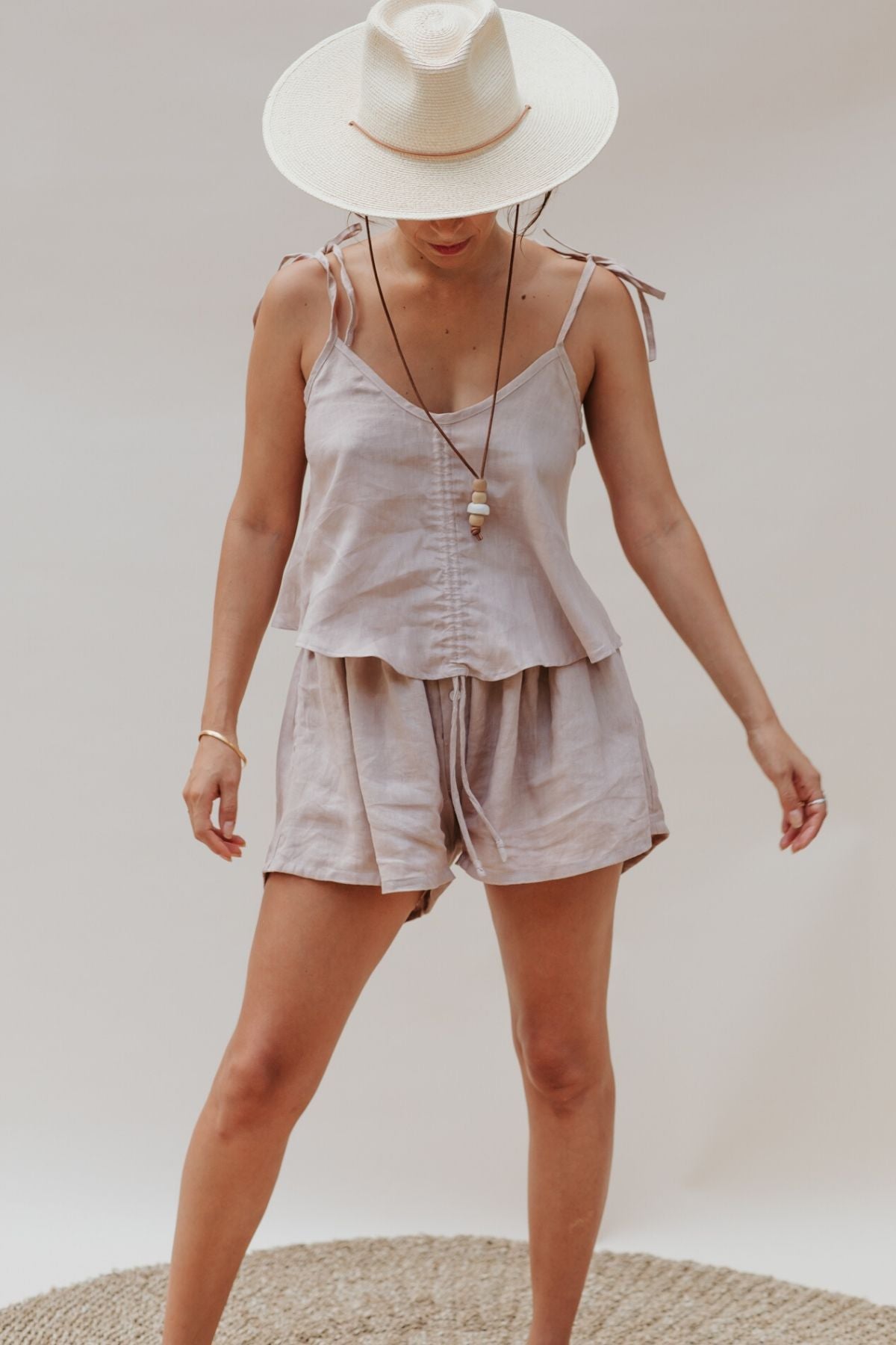 A person wearing a light beige camisole with adjustable straps and shorts stands on a round woven rug. They have a long necklace and a wide-brimmed hat. The simple, neutral background enhances the relaxed atmosphere, reminiscent of the elegance of Myrah Penaloza's Nidra Sleeper Top in Clay or the freshness of linen sheets on a sunny morning.