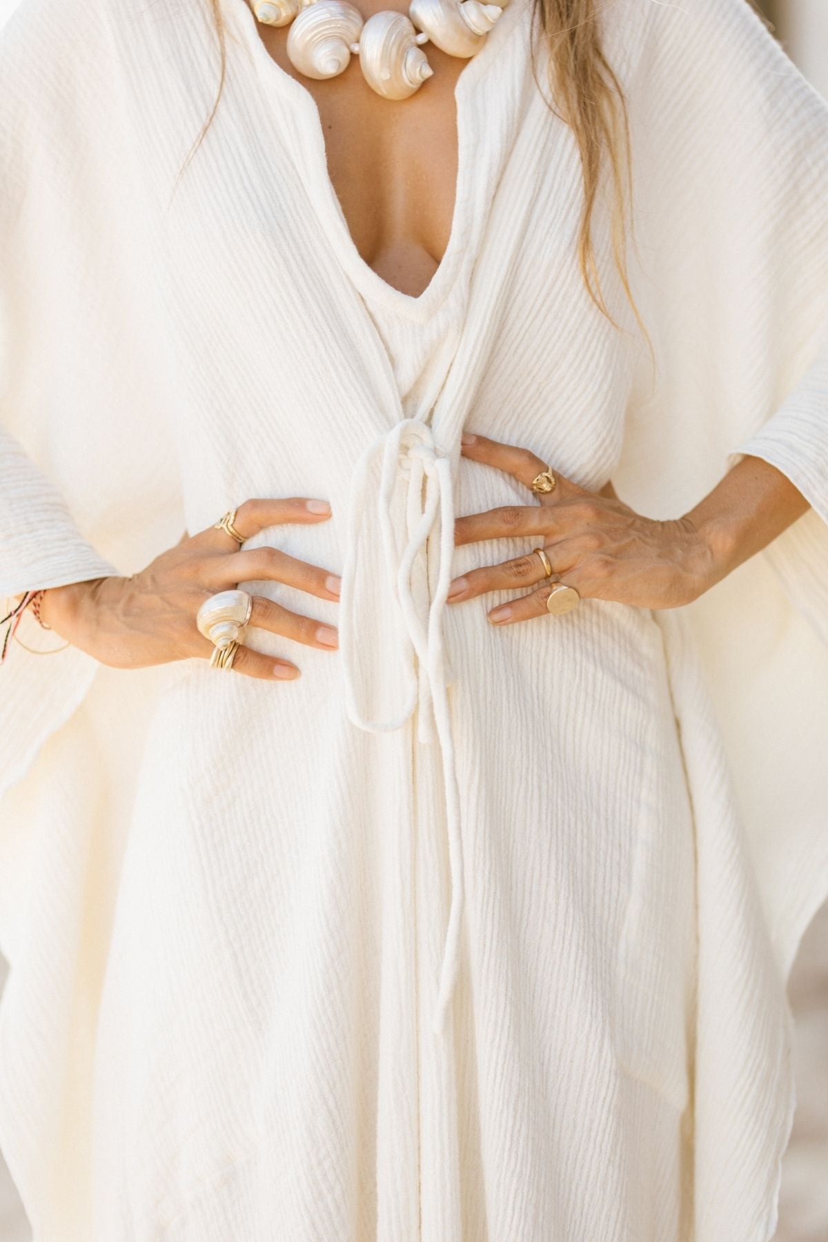 A woman stands with hands on her waist, wearing the Myrah Penaloza River Kaftan, made from 100% organic hemp and cotton. The kaftan features a textured white design, a deep V-neck, and a loose tie at the front. She accessorizes with several rings and a chunky necklace adorned with large white shells, all embodying an organic hemp style.