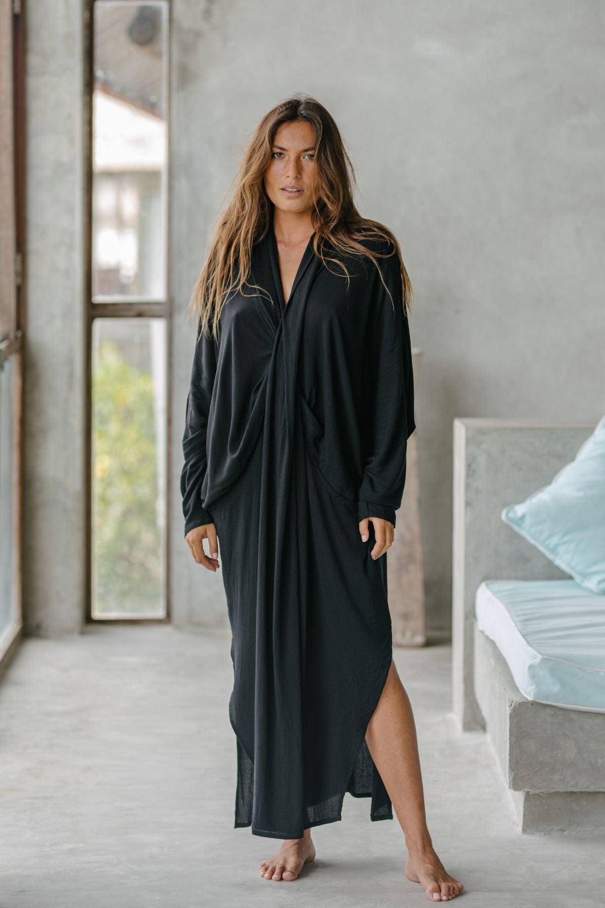 A woman with long hair stands barefoot on a concrete floor, wearing the loose-fitting Amma Gown - Dark Moon Edition by Myrah Penaloza. She is indoors, near large windows, with a light blue cushion on a nearby bench. The setting's minimalist design reflects intentional living ethos.