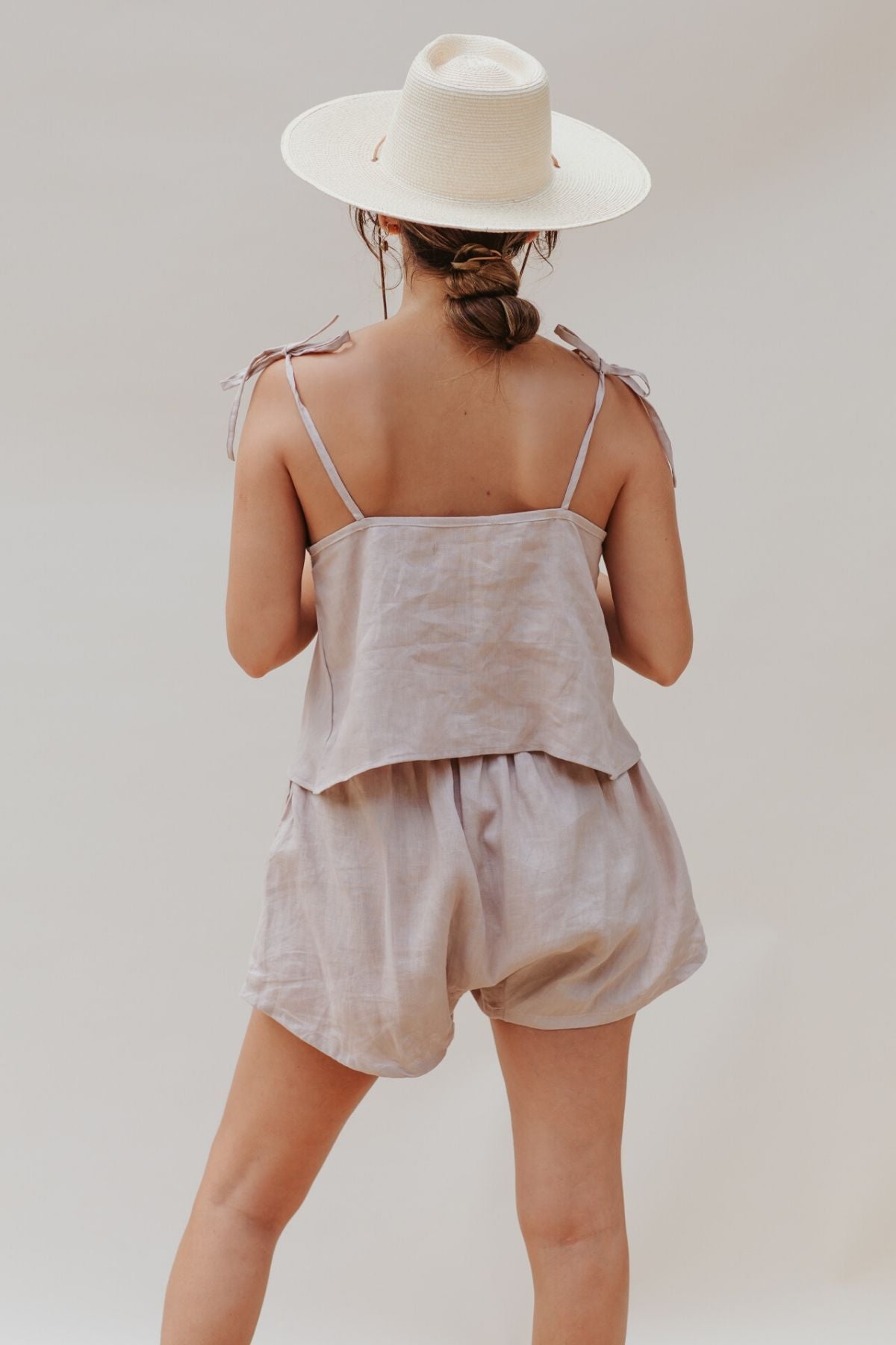 A woman wearing a beige sun hat and a matching Nidra Sleeper Top by Myrah Penaloza stands with her back to the camera. This lightweight, 100% linen clay-colored ensemble is perfect for relaxing moments akin to Nidra yoga. The neutral background enhances the serene vibe.