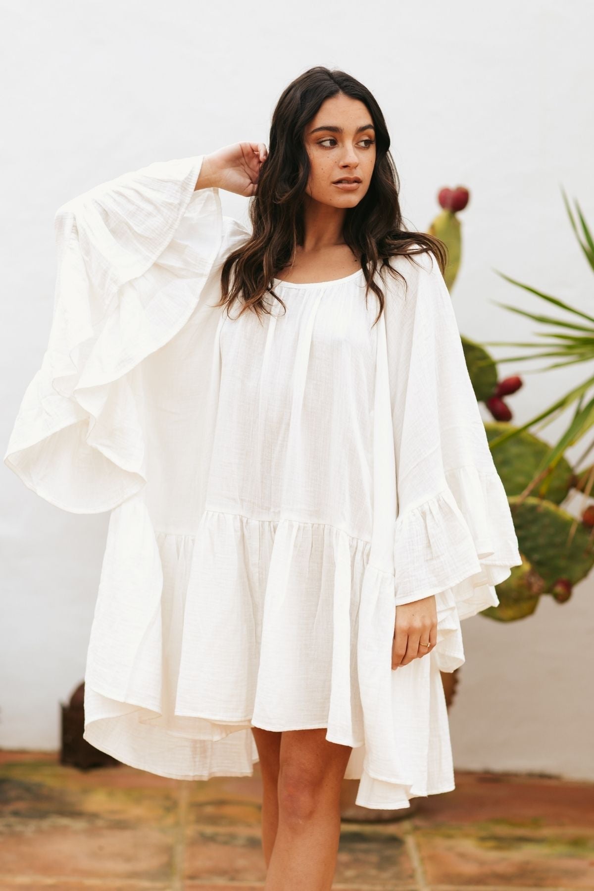 A woman stands outdoors, wearing the Brida Gown Short (WI) by Myrah Penaloza, characterized by its flowing white fabric with ruffled sleeves and hem that billow in a butterfly-like shape. The background features a white wall and green plants. She has long, wavy hair and her right hand is raised to adjust it, reminiscent of a character from Paulo Coelho's novels.