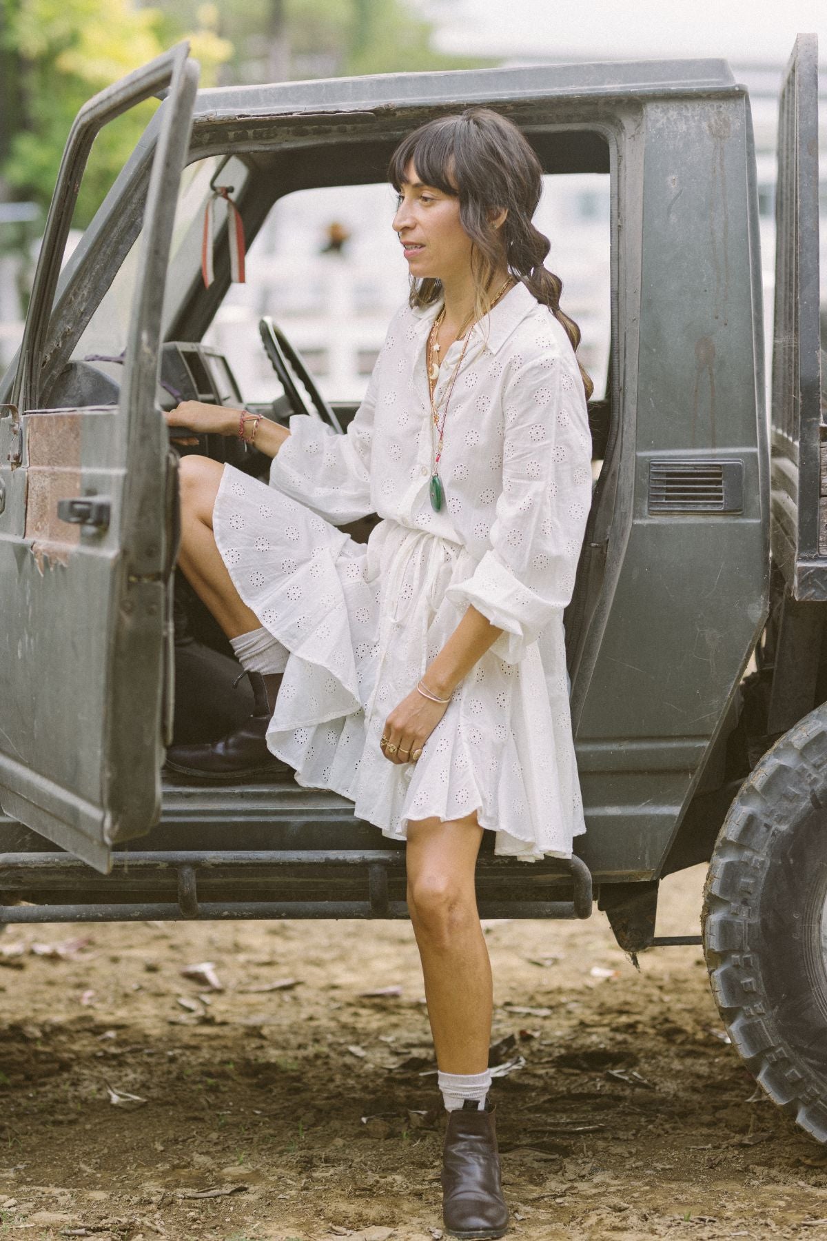 A woman wearing the La Rosa Dress by Myrah Penaloza, a vintage embroidered cotton piece, steps out of an off-road vehicle. She is holding the open door for support and looking away. The vehicle is parked on a dirt surface with greenery in the background. Her ensemble perfectly complements the scene, captured using Carbon Neutral Shipping practices.