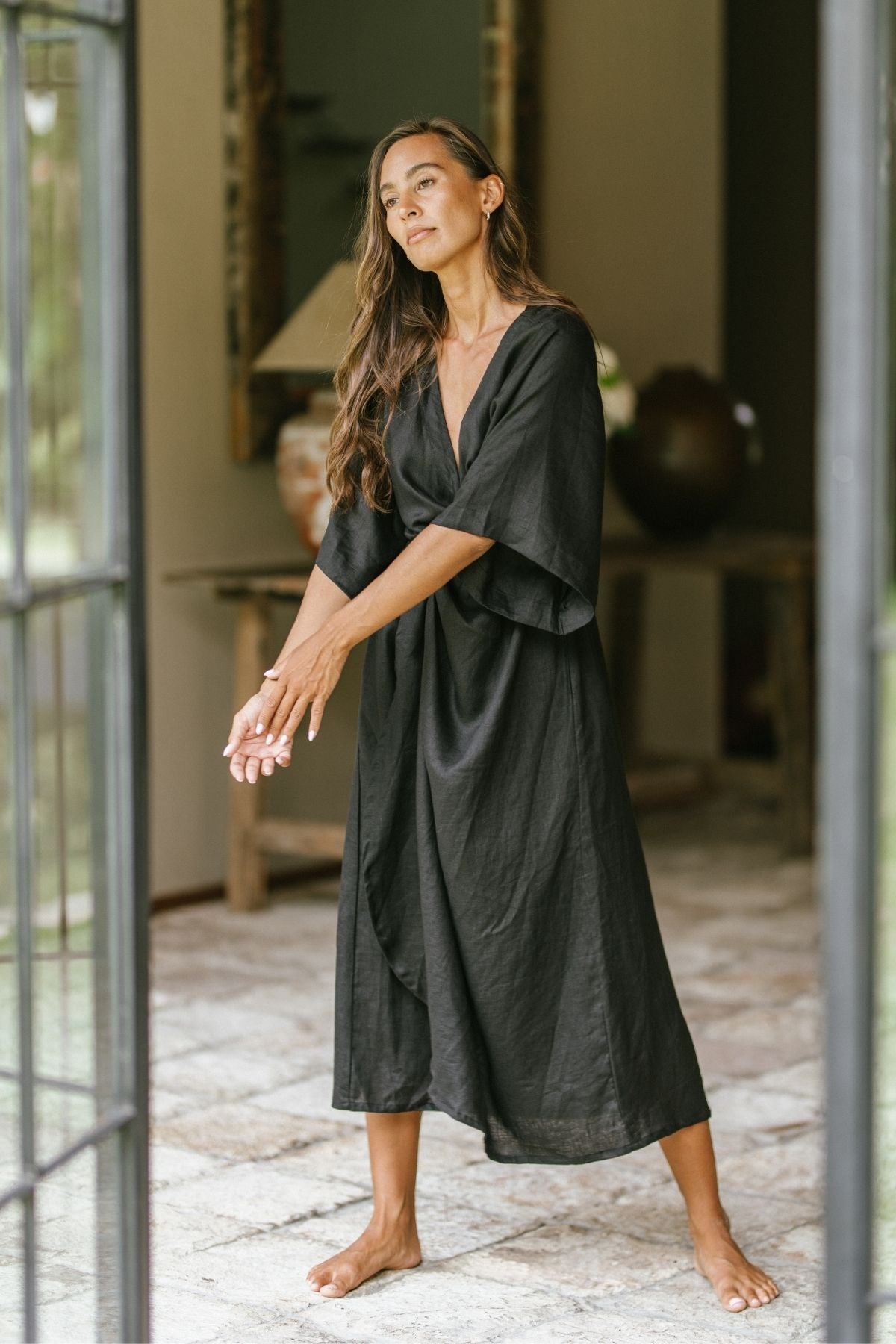 A woman stands barefoot in a doorway, wearing the Magdalena Gown by Myrah Penaloza, crafted from 100% linen in the Moonlight shade. Her long hair flows freely as she appears relaxed with her arms slightly raised. The background reveals a room adorned with a table and decorative items.