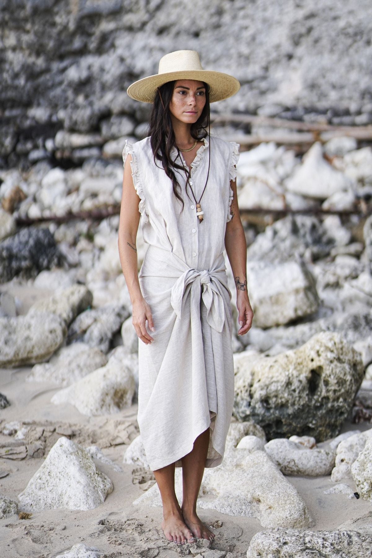 Standing barefoot on a rocky beach, a person with long hair dons a wide-brimmed straw hat and The Madu Linen Wrap Dress by Myrah Penaloza. The sleeveless, light linen dress is elegantly tied at the waist, while the surrounding rocks and sand accentuate their peaceful presence.