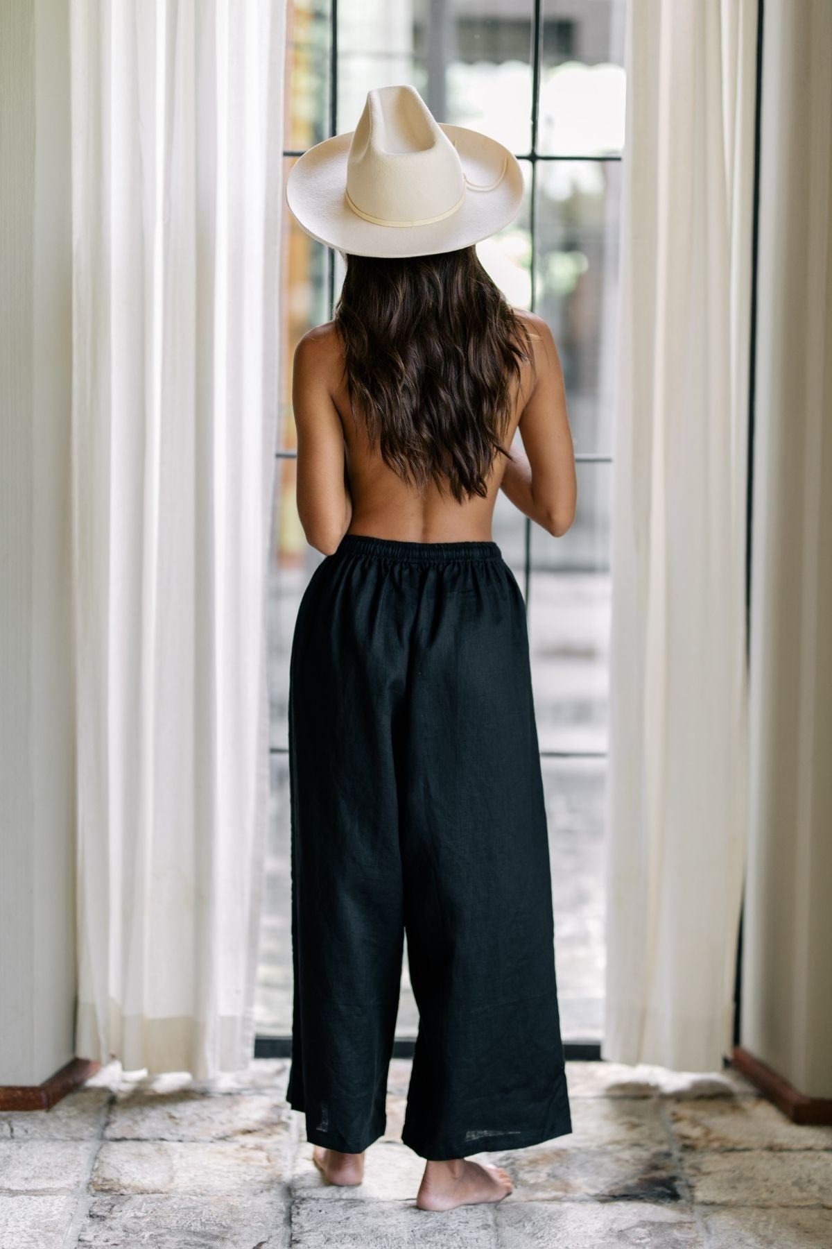 A person with long brown hair stands barefoot and topless, wearing the dark moon black Grace Pant by Myrah Penaloza, which is crafted from 100% linen. They also wear a wide-brimmed hat as they face away from the camera, gazing out through sheer white curtains and glass doors behind them.