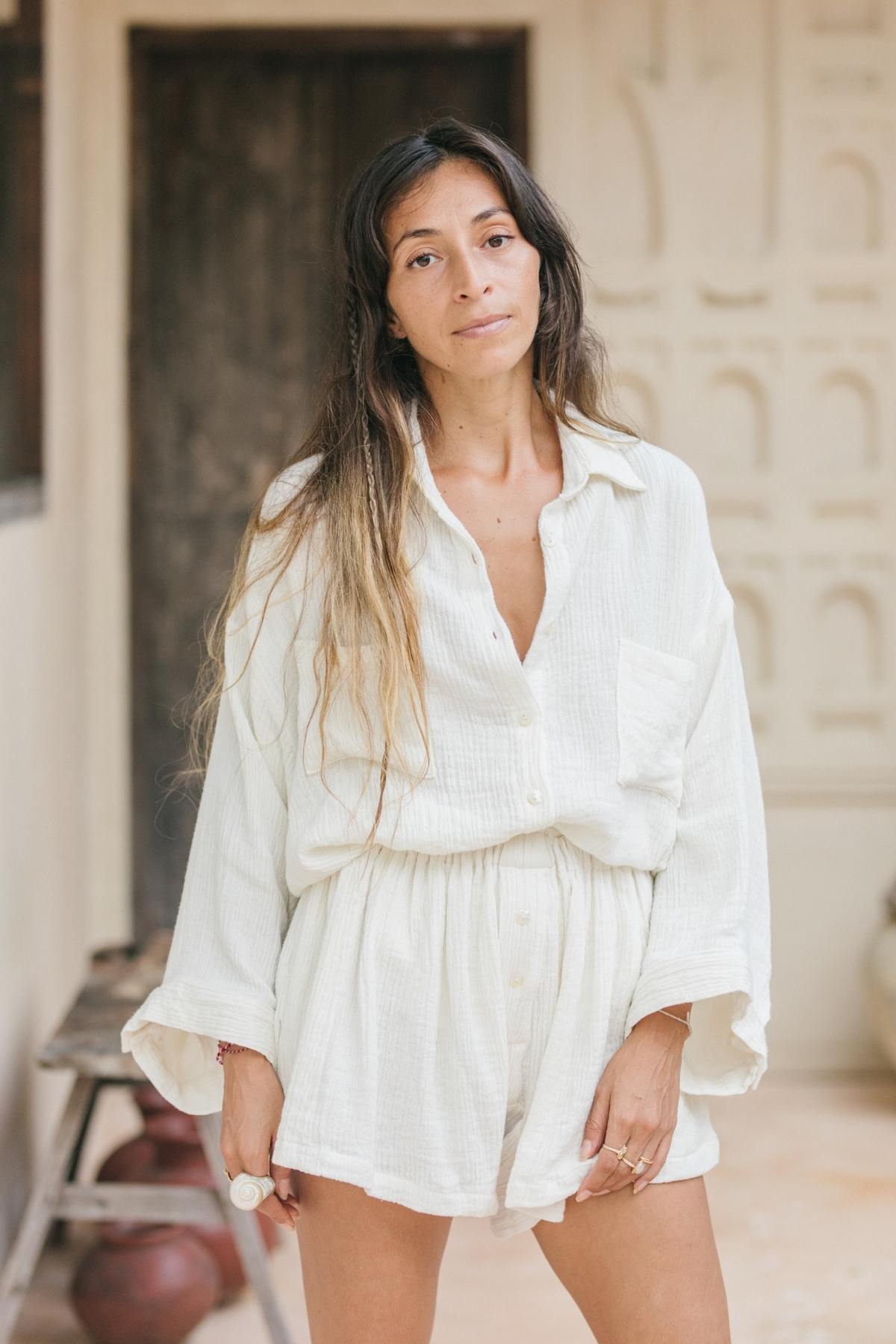 In a serene and airy setting, a person with long hair stands in a relaxed pose, dressed in the Nidra Button Down Top + Nidra Short Set by Myrah Penaloza, crafted from organic cotton and hemp. The background showcases a textured wall alongside a wooden door.