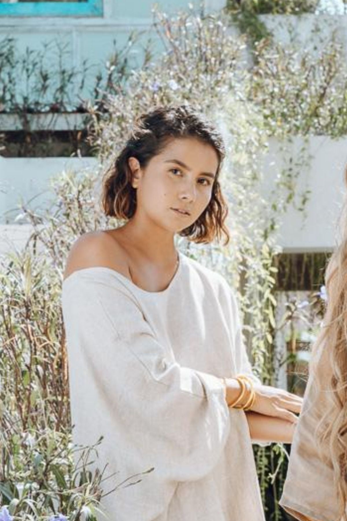 A person with shoulder-length hair is standing outdoors, dressed in the Myrah Penaloza Butterfly Tunic, which is loose and light-colored. The background showcases greenery and a building, creating a tranquil and natural atmosphere—an everyday piece ideal for stylishly embracing nature.