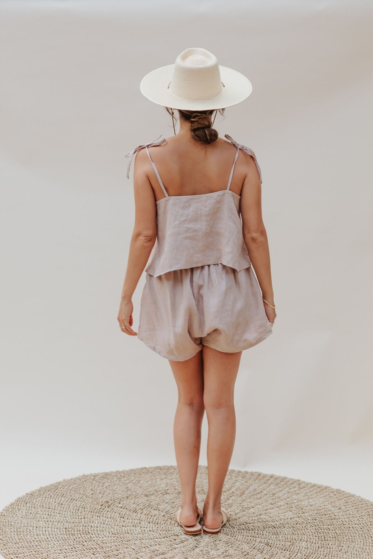 A woman stands on a woven rug, facing away. She wears a light beige sun hat, a sleeveless Nidra Sleeper Top by Myrah Penaloza in clay color made of 100% linen, with adjustable straps and matching shorts. The background is plain and neutral, creating a minimalist setting.