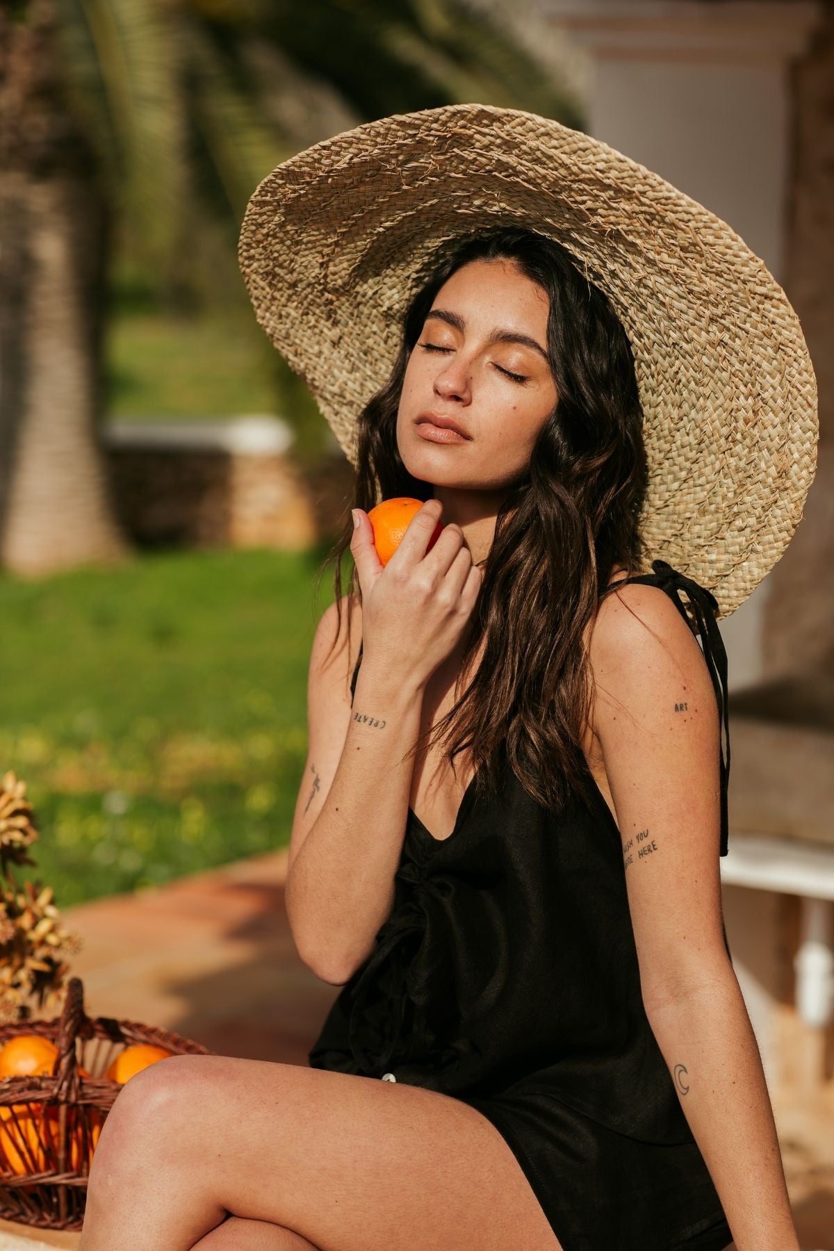 A woman wearing a Nidra Sleeper Top in clay, designed by Myrah Penaloza, pairs her attire with a wide-brimmed straw hat as she relaxes outdoors. She holds an orange near her face with closed eyes, sitting beside a basket of oranges against the serene backdrop of green grass and palm trees.