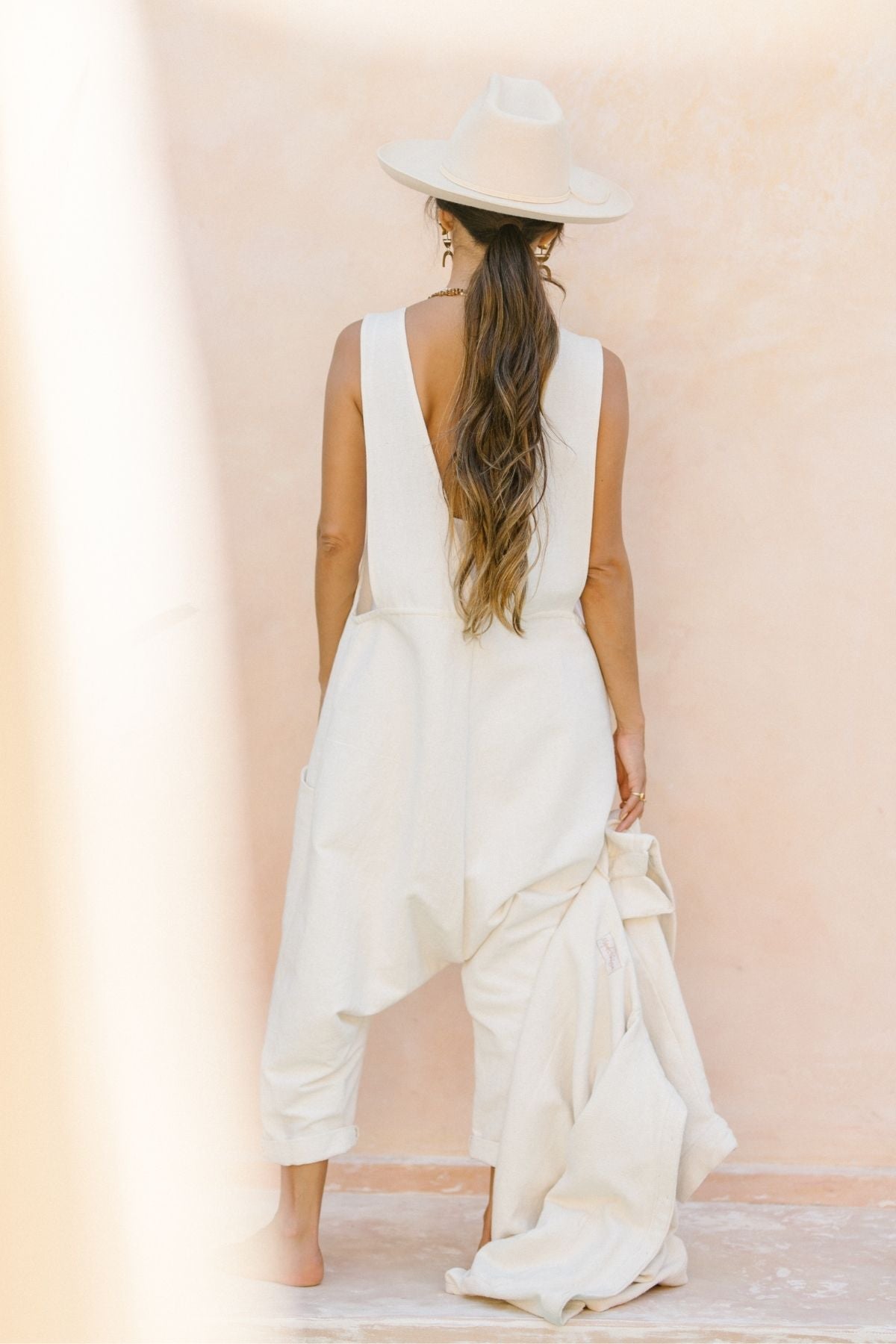 With long hair cascading down, a person stands with their back to the camera, exuding a vintage look in the Freya Overalls Artist Edition (Canvas Playsuit) by Myrah Penaloza. Their wide-brimmed hat complements the white sleeveless jumpsuit as they hold a matching coat against a light, textured wall.